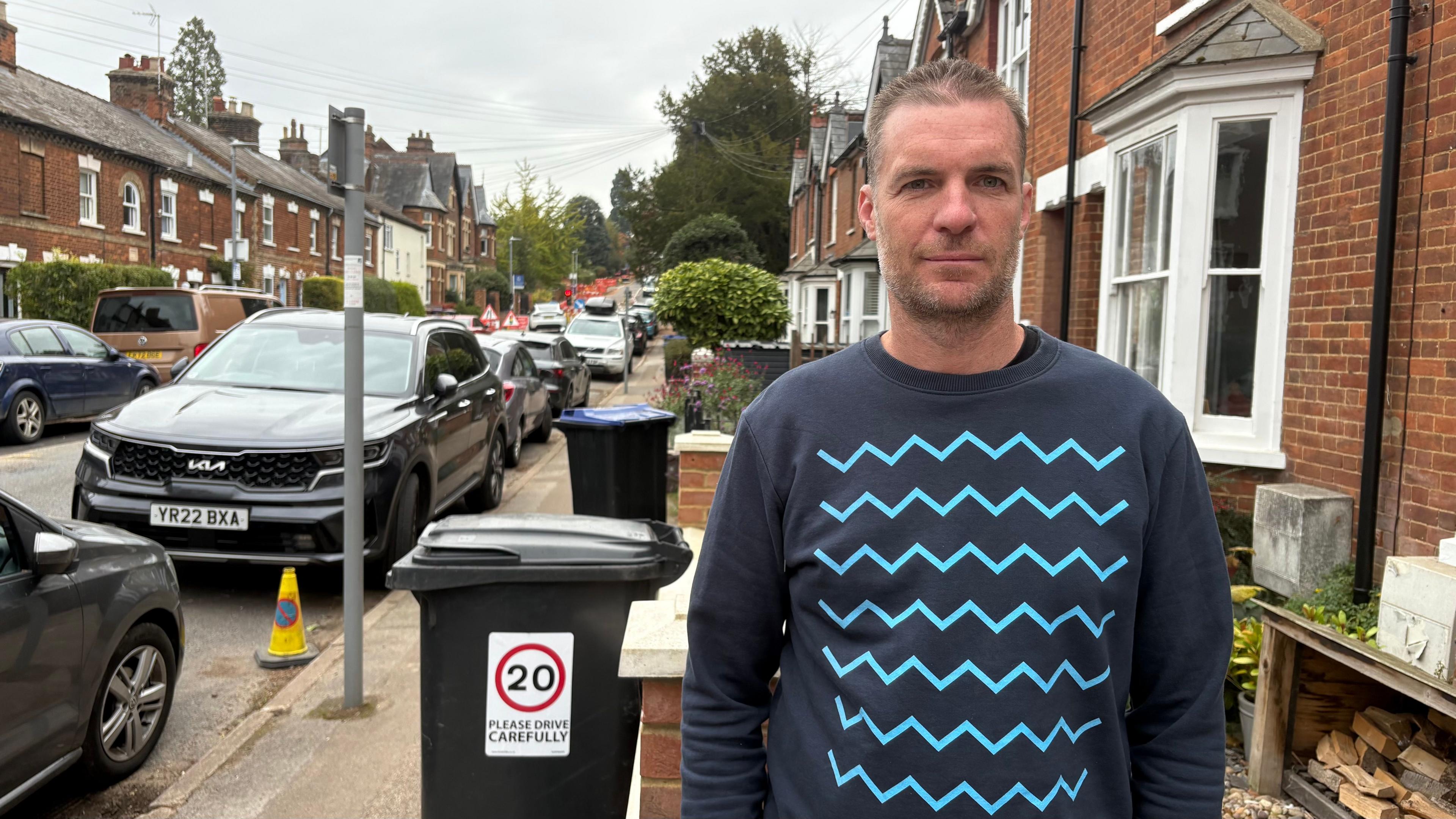 Jodie Trowsdale with short dark hair, looking at the camera and wearing a dark blue sweater with a blue wave pattern on the front. There is a wheelie bin with a 20mph sticker on the side. There are cars parked on either side of the street and old-style houses on both sides.