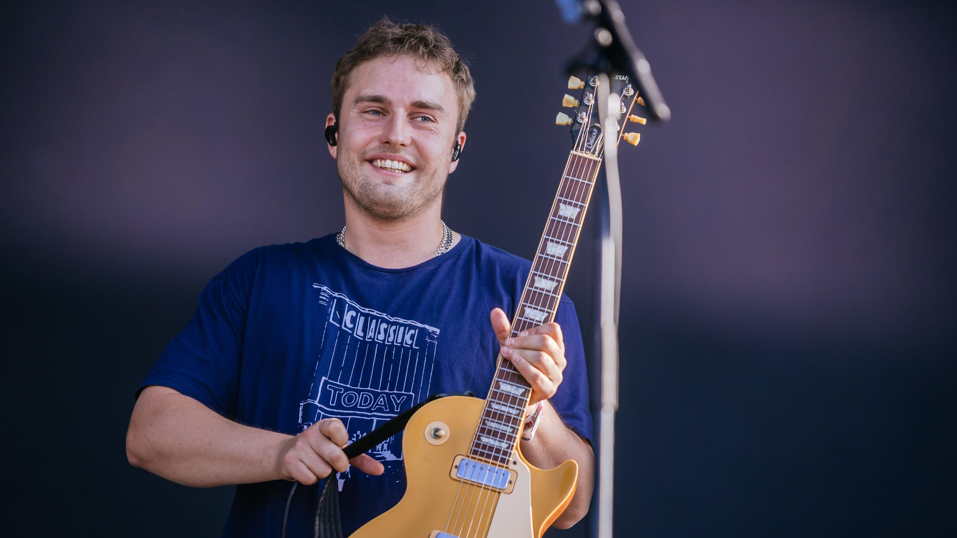 Sam Fender performing onstage during Southside Festival at Take-off Gewerbepark on 22 June, 2025 in Neuhausen, Germany. He has short, curly brown hair and stubble on his face. He is smiling and holding up a yellow electric guitar. He is weaing a navy t-shirt with a "Classic Today" logo and has a set of black earbuds in his ears.