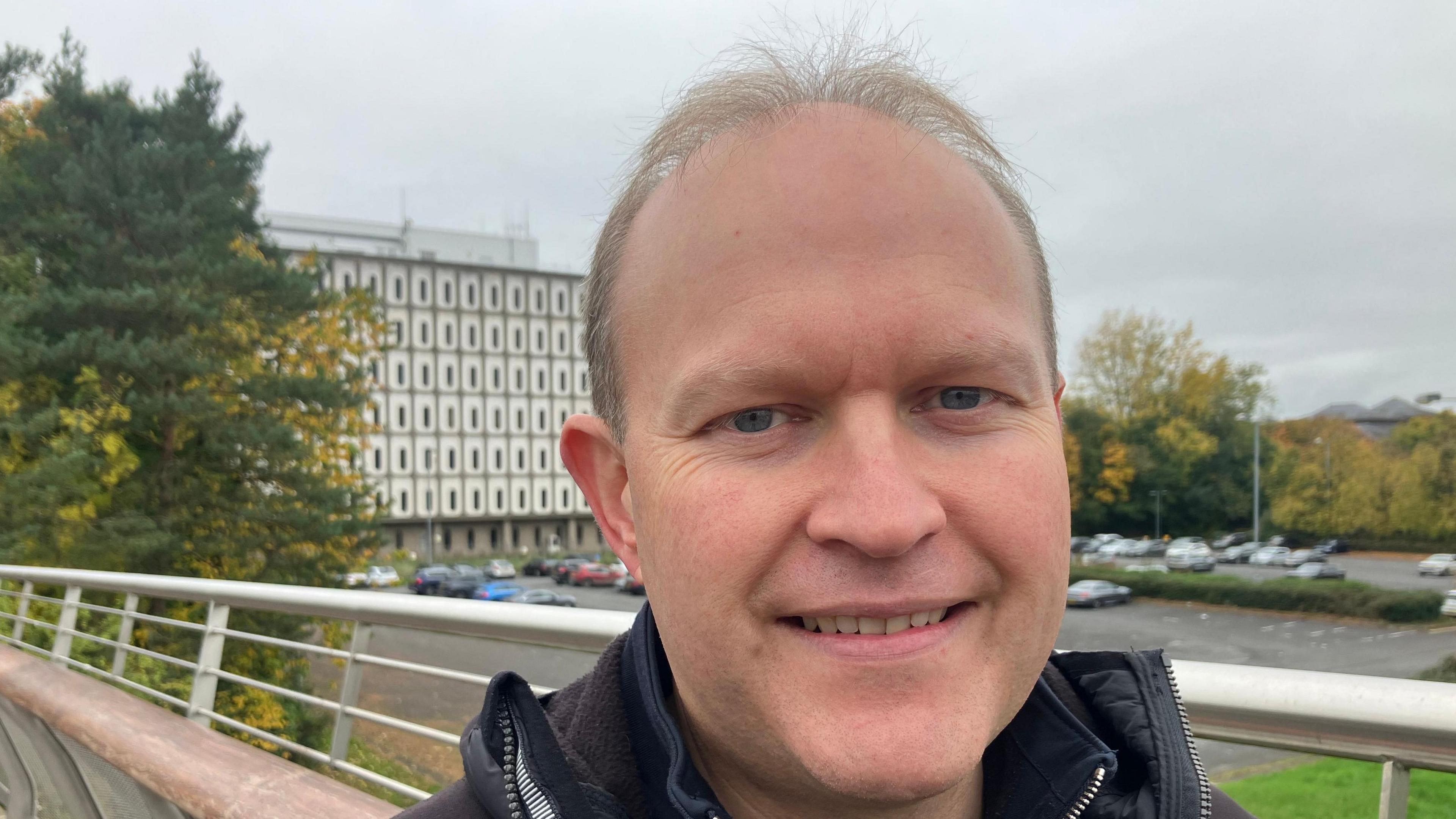 Gareth Wilson pictured at a bridge leading to Marlborough House. He has short light hair and is wearing a black coat. He is smiling at the camera.