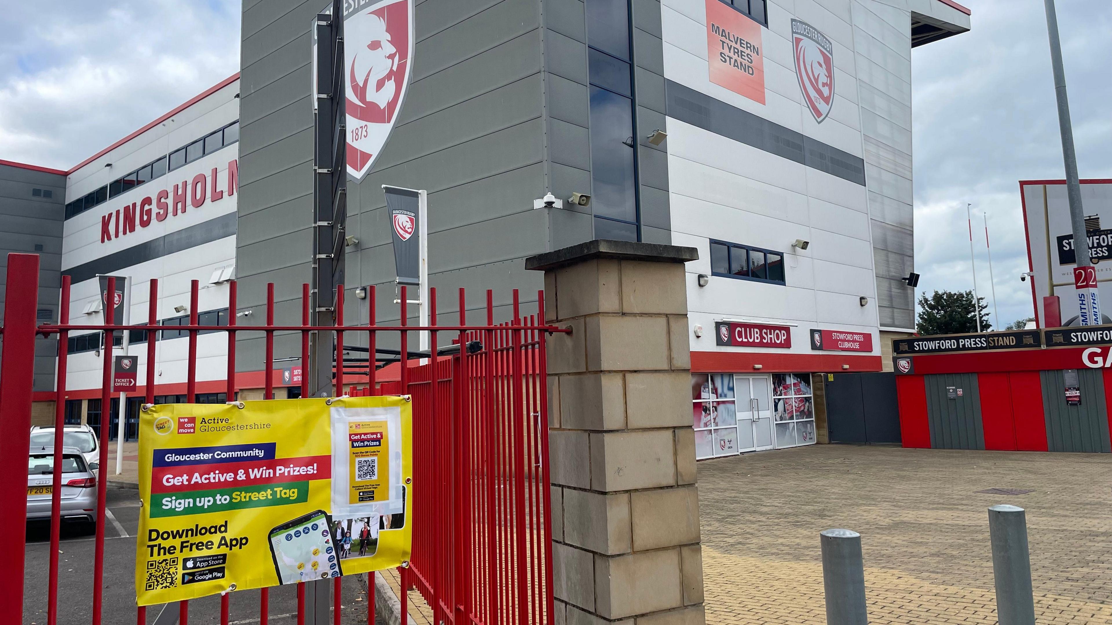 Image shows a yellow poster with the Street Tag QR code that has been tied to red gates outside of Kingsholm Stadium.