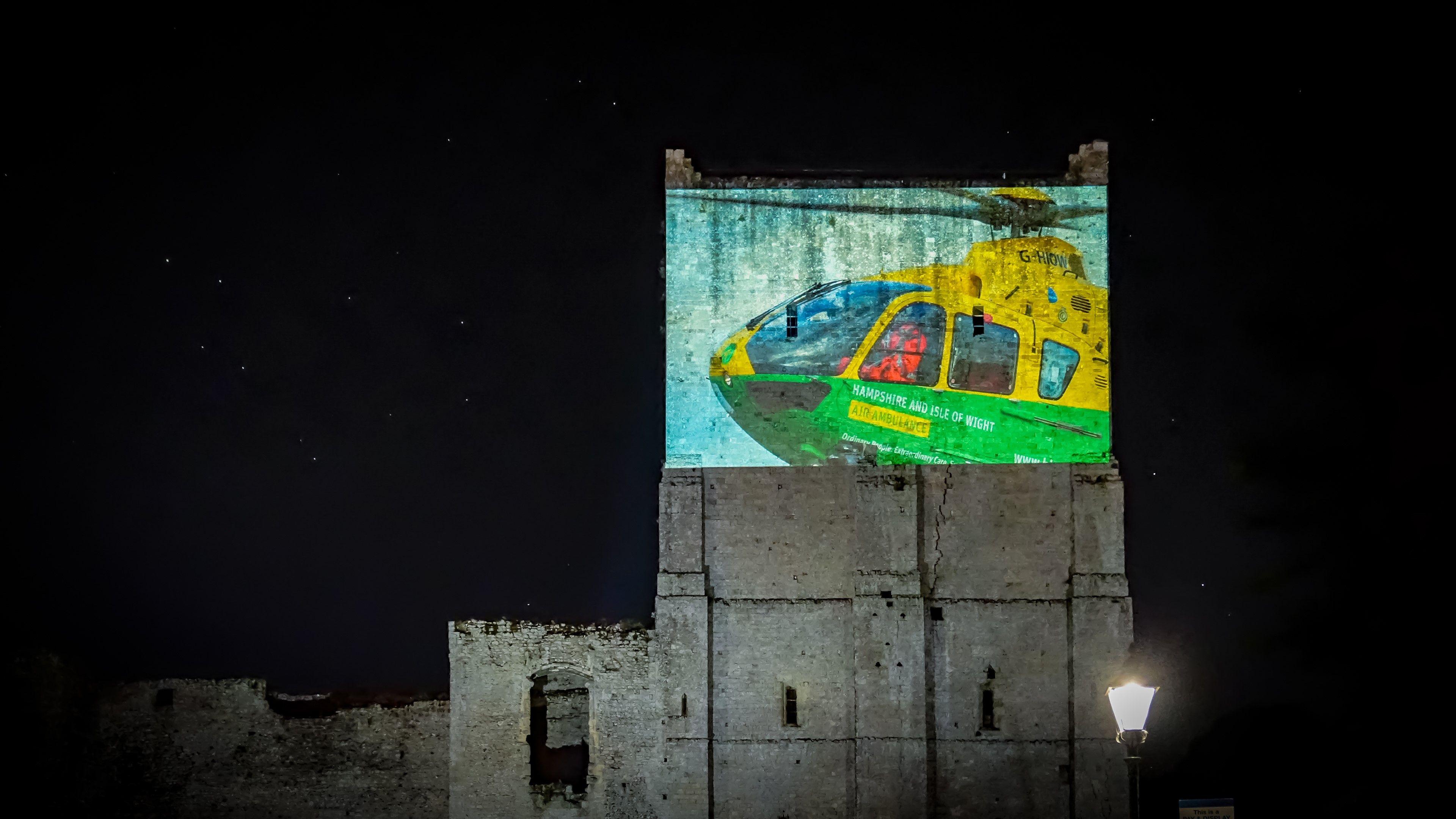 An old castle building at night time. It is tall and in the top half, an image of a green and yellow helicopter is projected on to it.