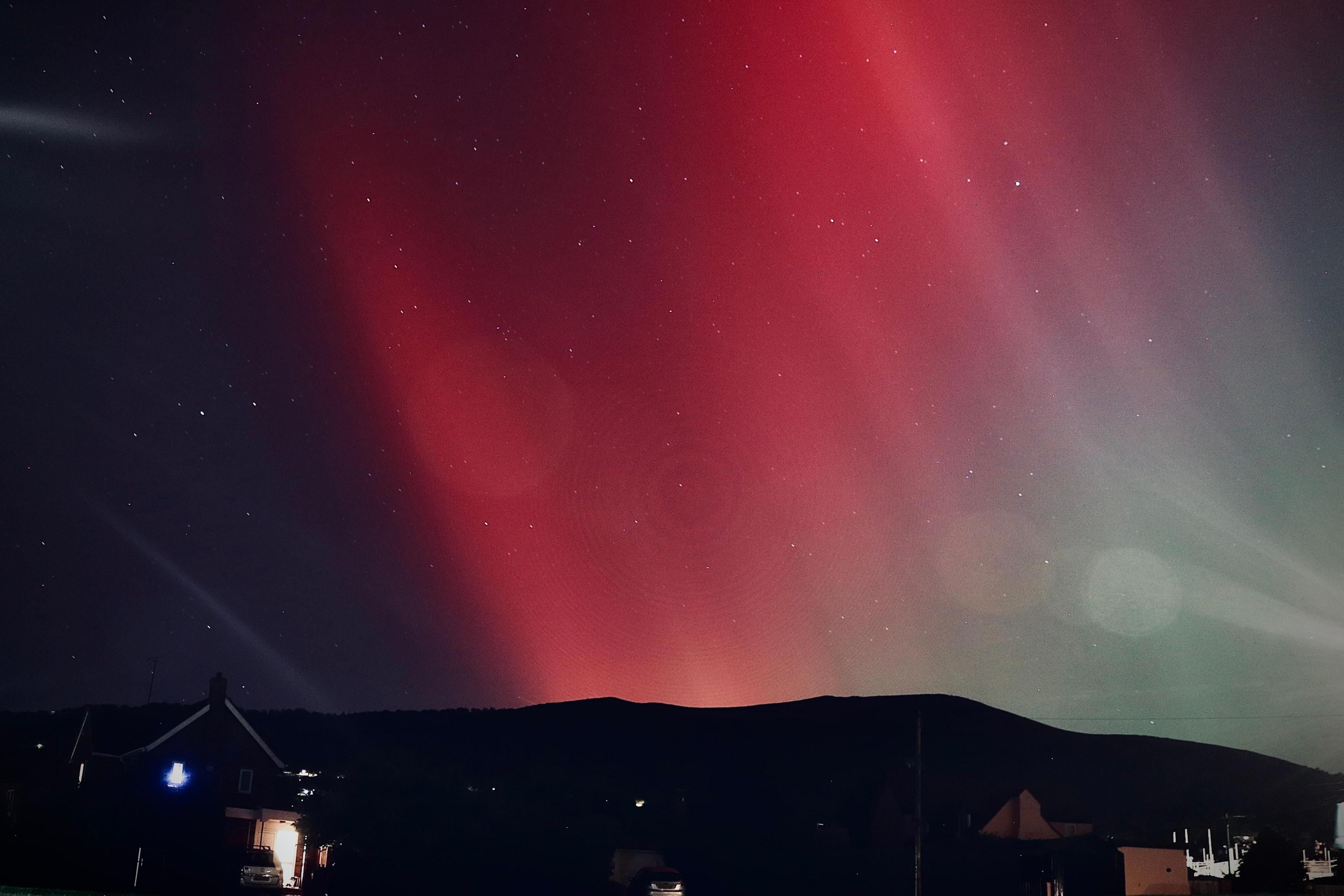 Sheets of red run diagonally through the sky, with a house and hillside in the foreground