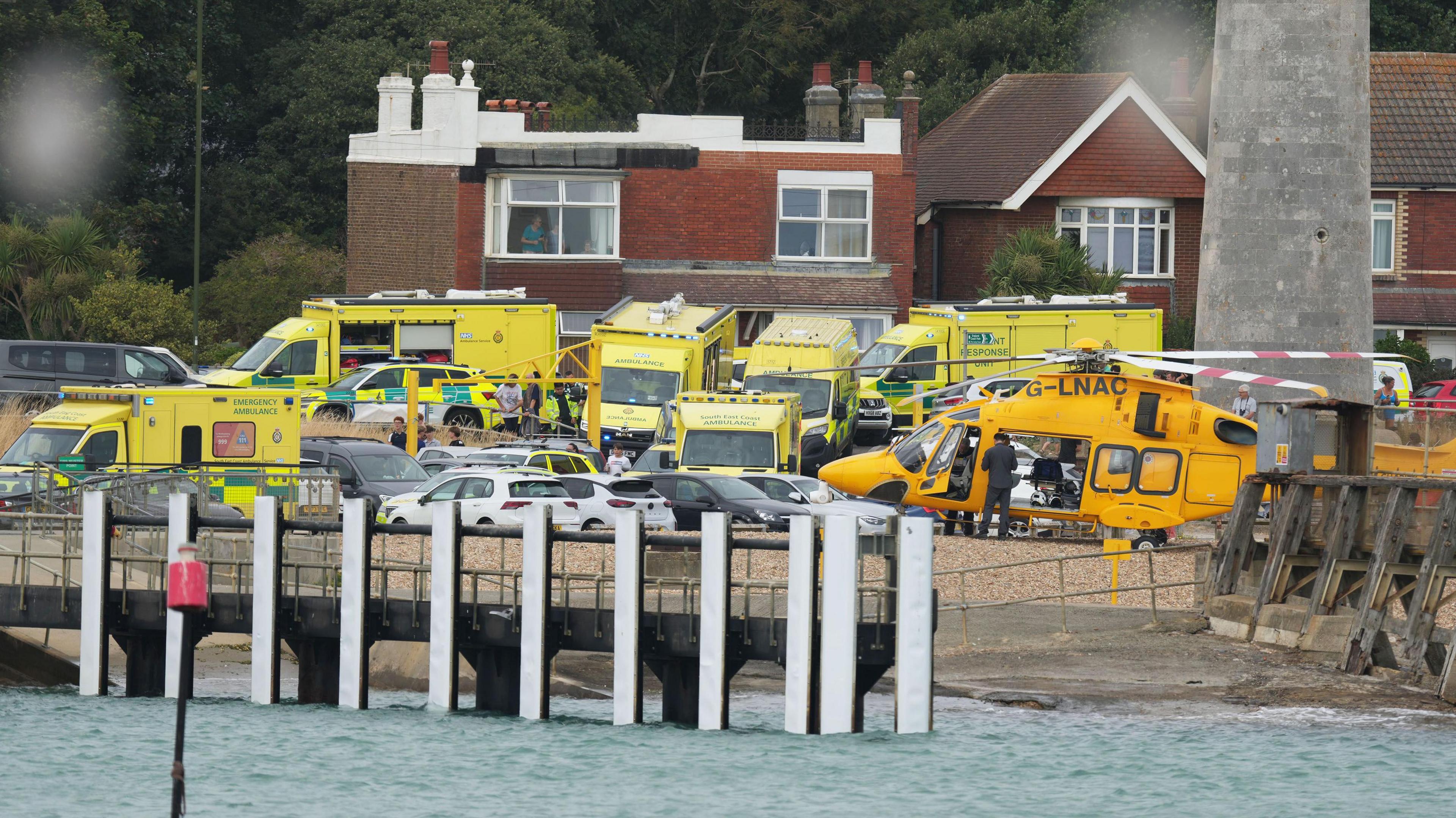 Multiple ambulances and and air ambulance can be seen behind a handful of parked cars at the Shioreham waterfront. The sea can be seen in the foreground, with breakwaters. Behind the ambulances are houses. Several people can be made out amound the emergency vehicles