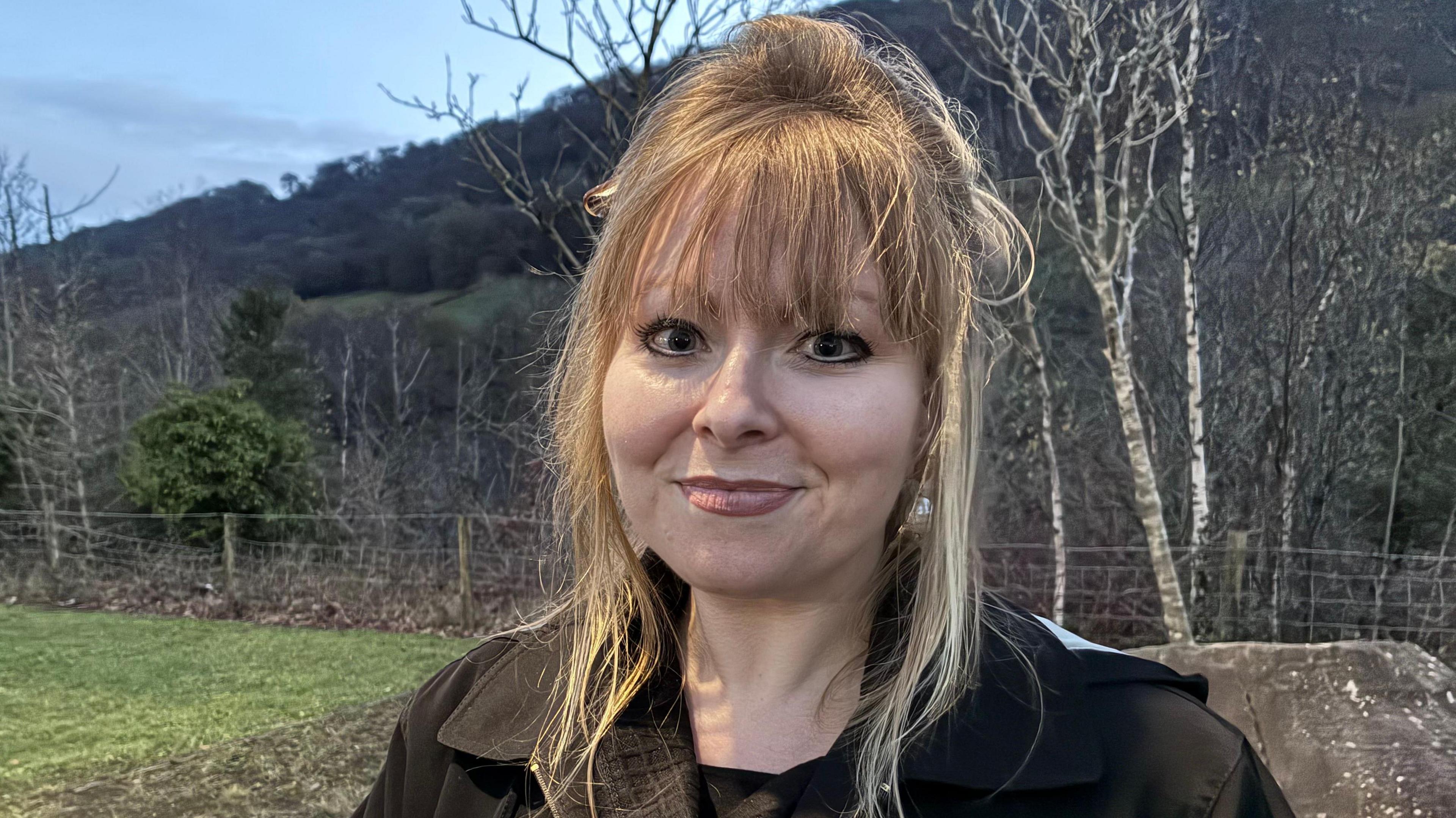 Delyth Jewell yn edrych syth at y camera. Mae hi tu allan gyda golygfa o goed ar fryn tu ol iddi. Mae hi'n gwisgo cot cynnes.