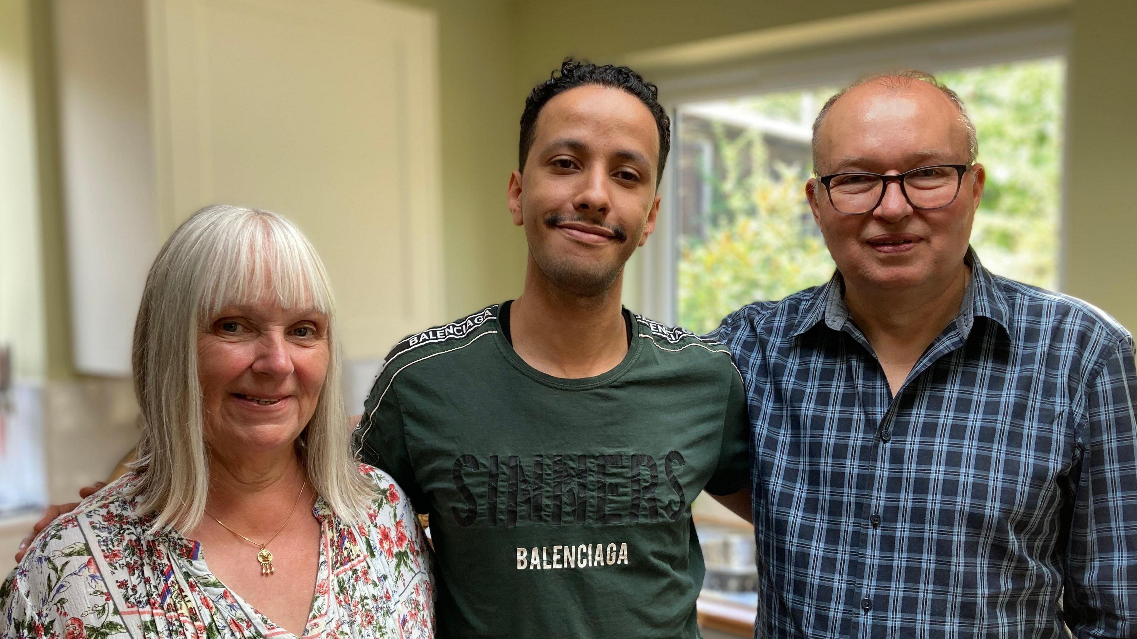 Anita is on the left of the picture. She has shoulder length white hair and is wearing a flowery blouse and a gold necklace. Aziz is in the middle and has short black hair and a green T-shirt that says "Sinners: Baleciaga". Steve is on the right of the picture and he is wearing a checked blue and dark blue buttoned shirt and he has dark glasses. He is balding but has a bit of short hair on his head.