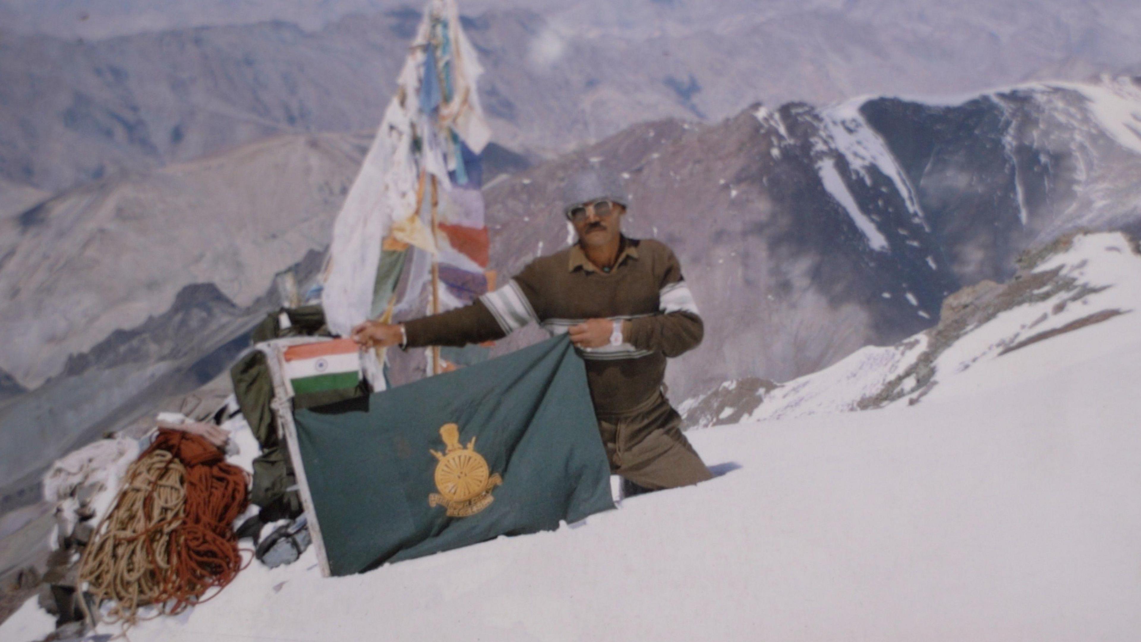 A man wearing protective clothing on a snow-capped mountain peak holds a green flag, with a smaller Indian flag above it.   
