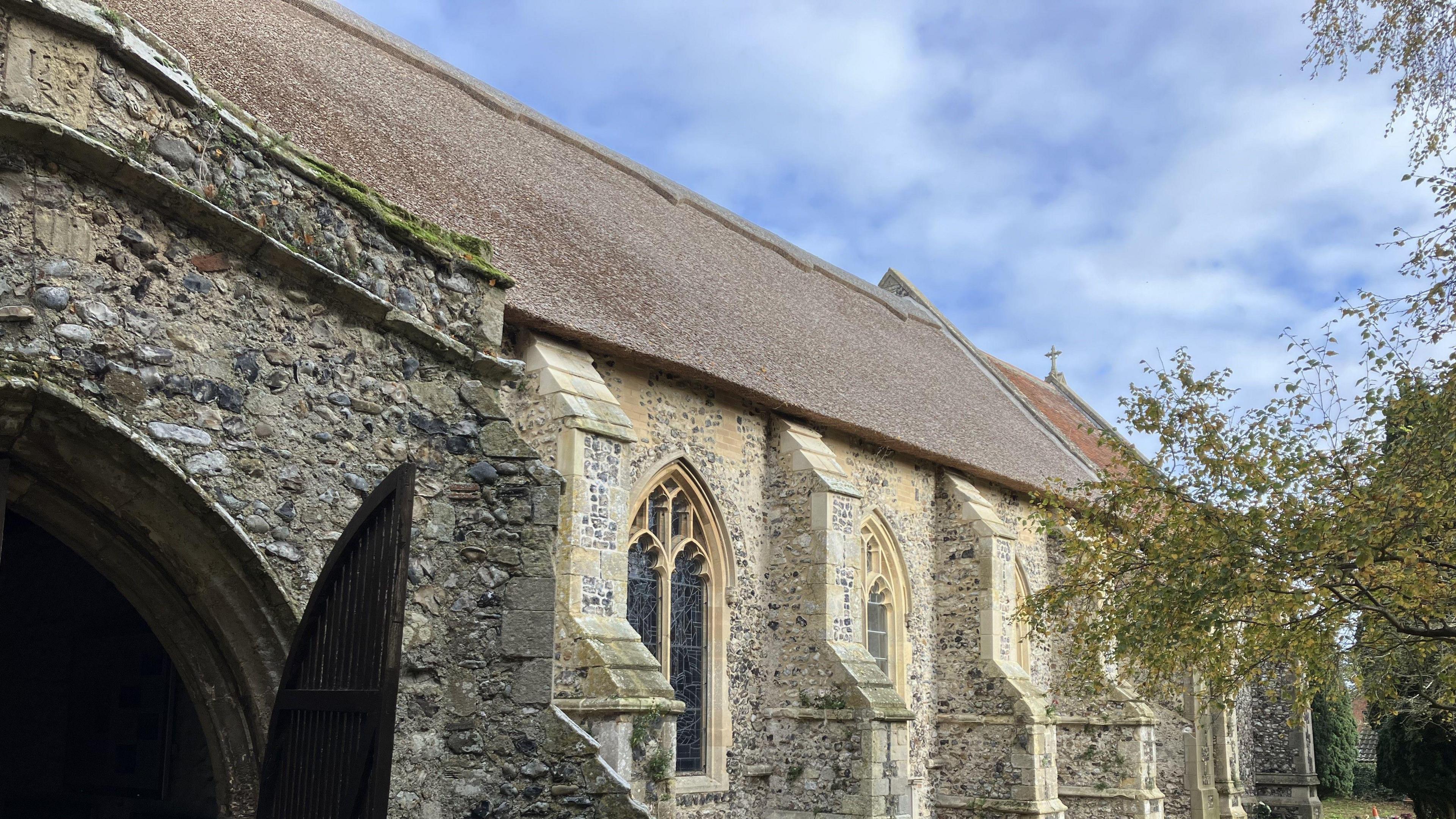 A side view of the church showing the opened door of the south porch, the stone buttresses and the newly-repaired thatched roof