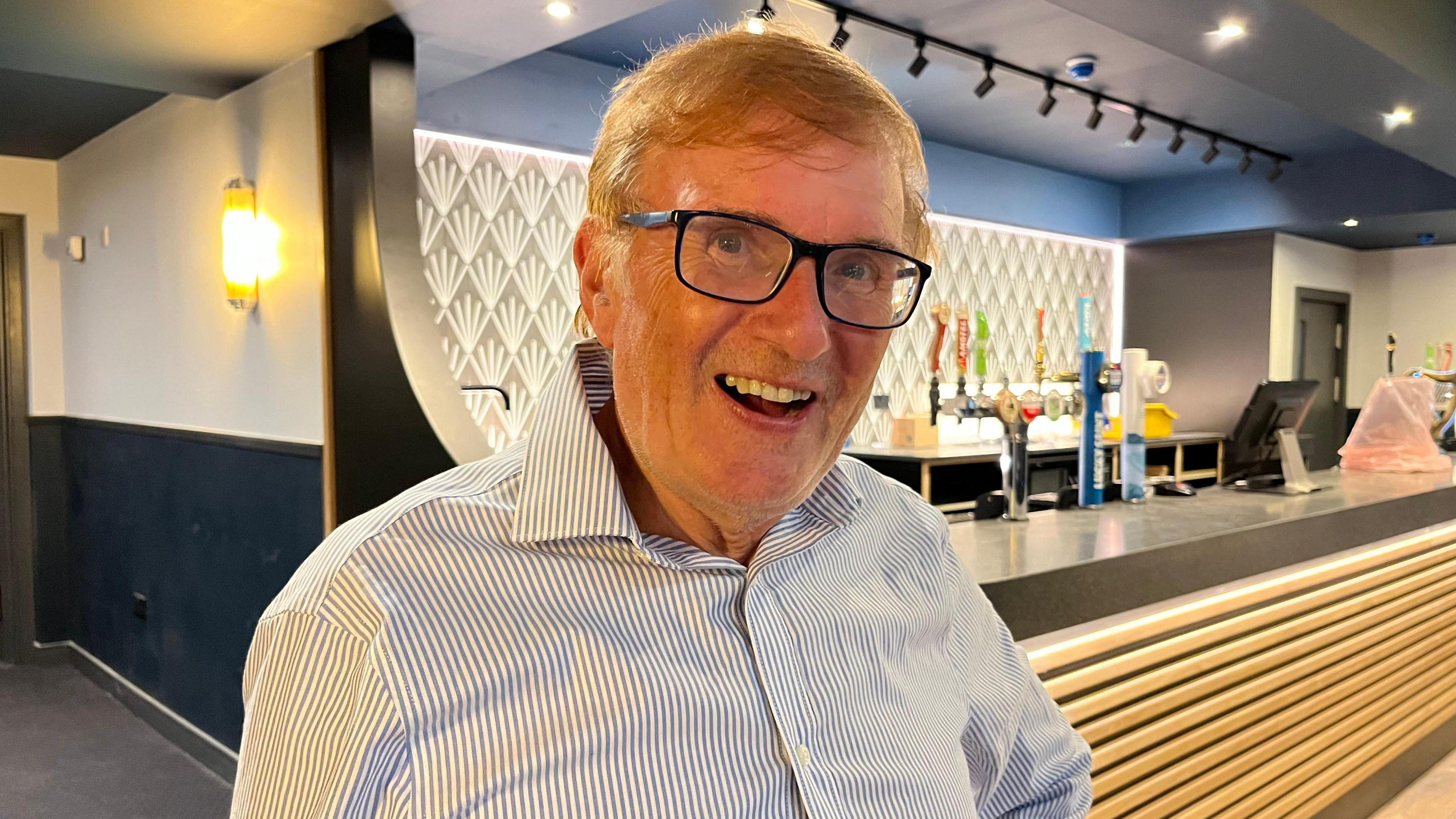 A smiling Geoff Greaves stands in front of the bar in the Ritz. It has a 1930s style light and a curved edge to the bar. Mr Greaves is wearing glasses and a striped shirt.