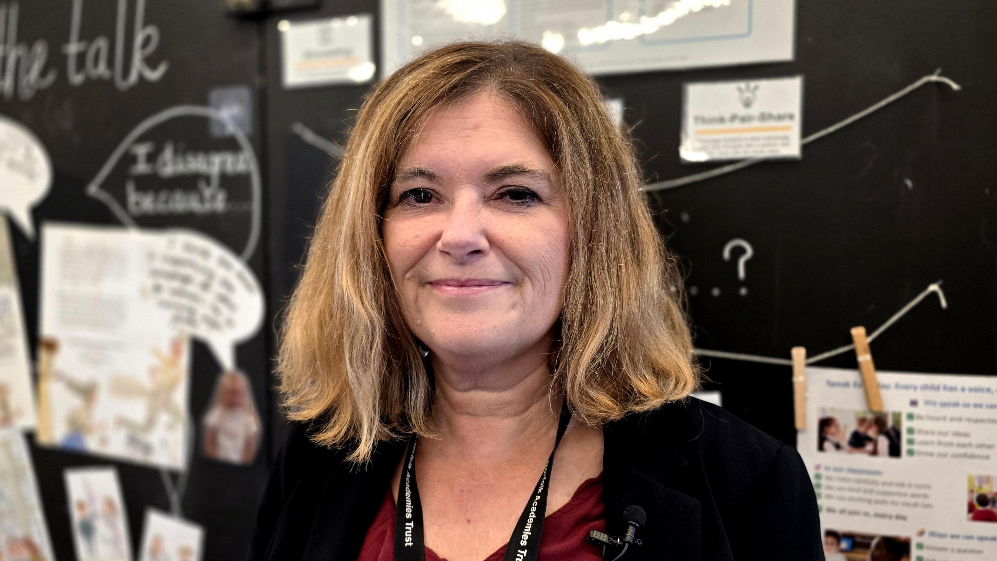 Louise Jackson is smiling at the camera and standing in front of boards with various bits of school work on. She's wearing a dark jacket over a crimson top and is also wearing a lanyard.