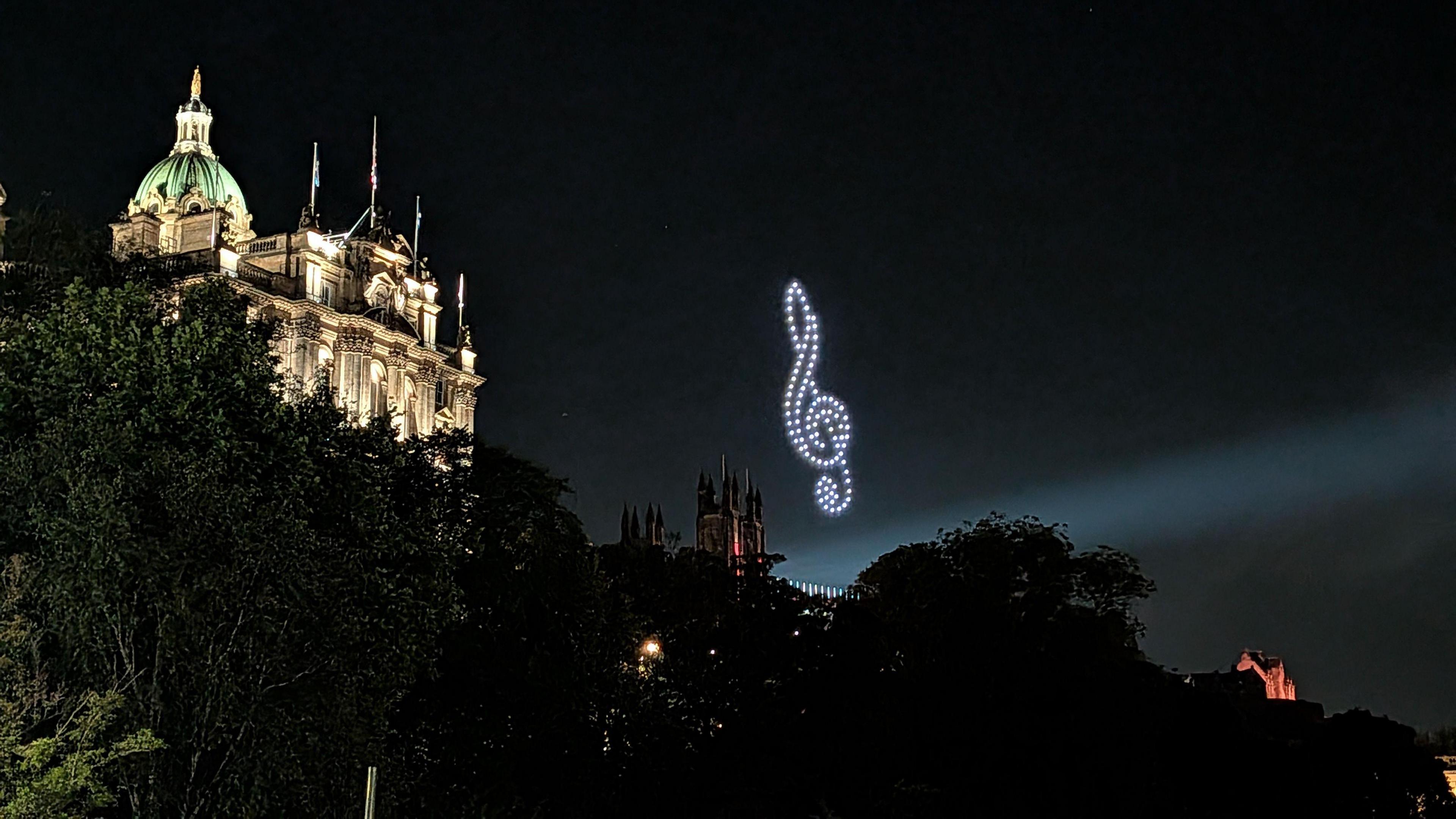 Edinburgh monuments lit up at night. Drones make the shape of a treble clef in the sky.