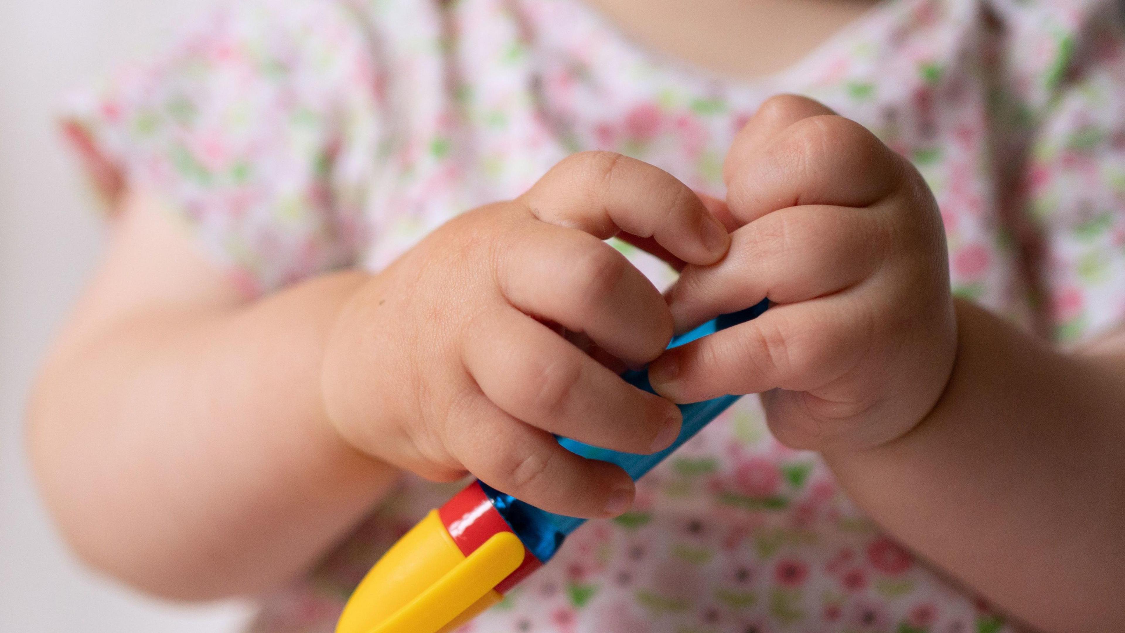 Close up of a young child's hands - the child is wearing a pink flowery top and holding a plastic blue, red and yellow pen