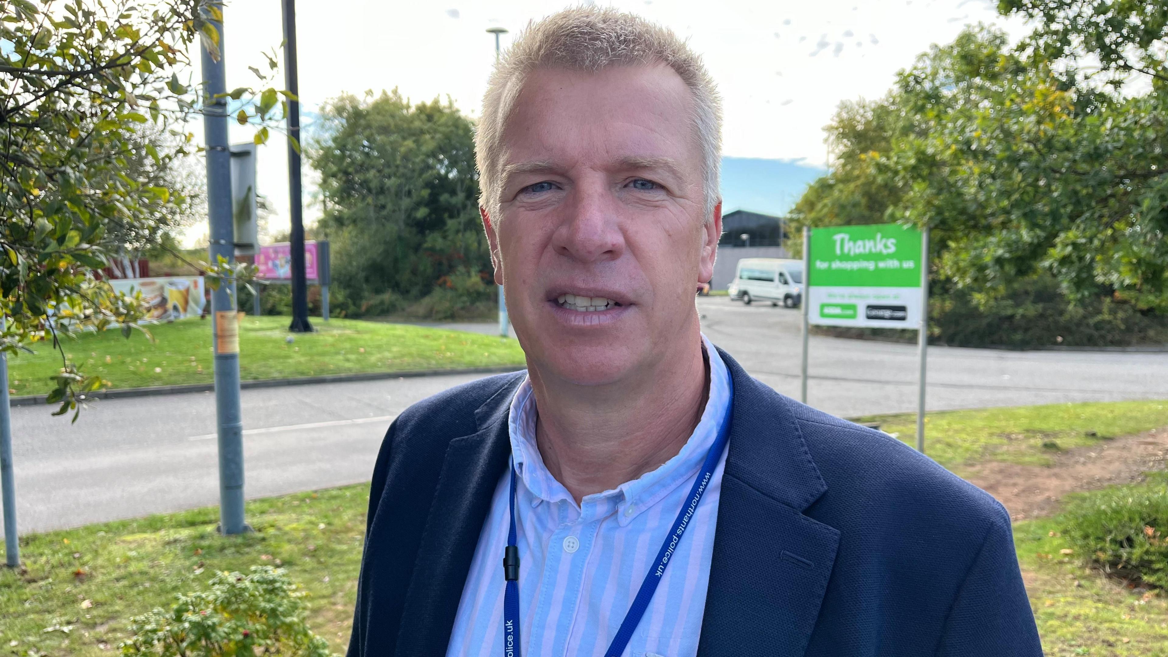 Neil Cowley in blue suit with short blonde hair, standing in front of trees and a road