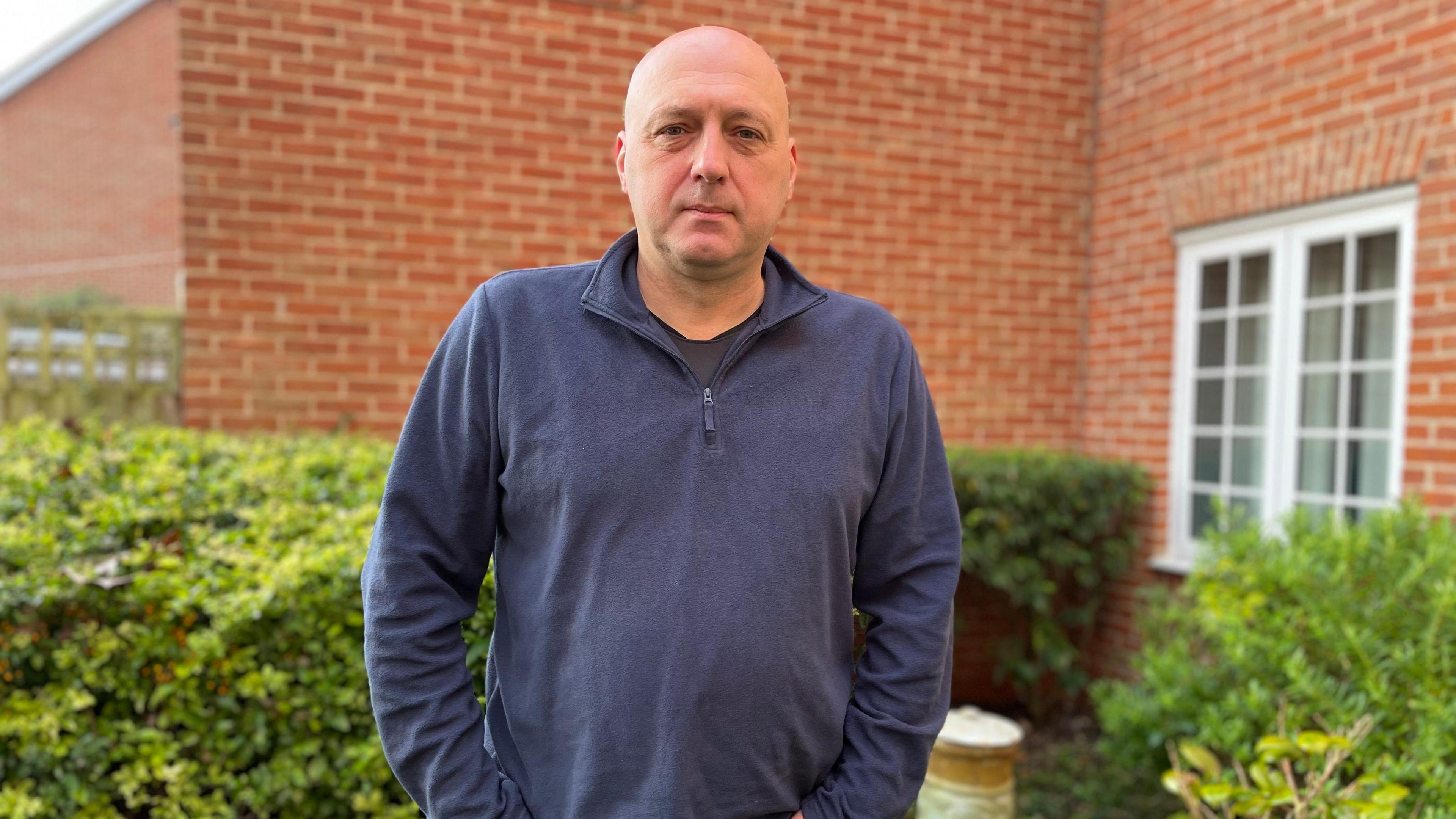 Colin Phillips stands outside a modern brick-built house, with a hedge and shrubs behind him. There is a white window in the side of the house. He is bald and wearing a navy blue open-collared jersey top.