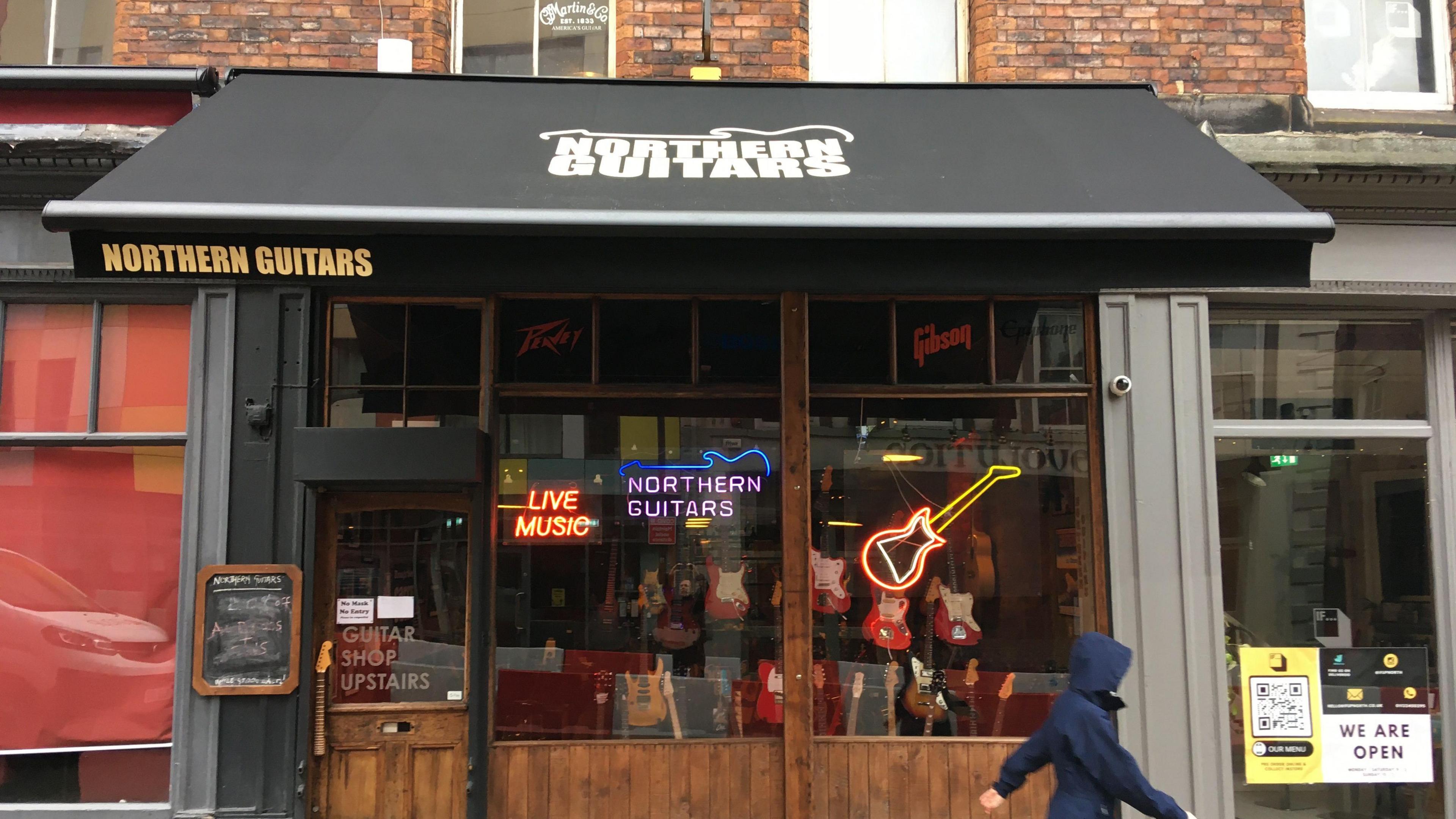 Northern Guitars shopfront. A black awning over a guitar shop with a dark wooden frontage. A person walks past in a raincoat.