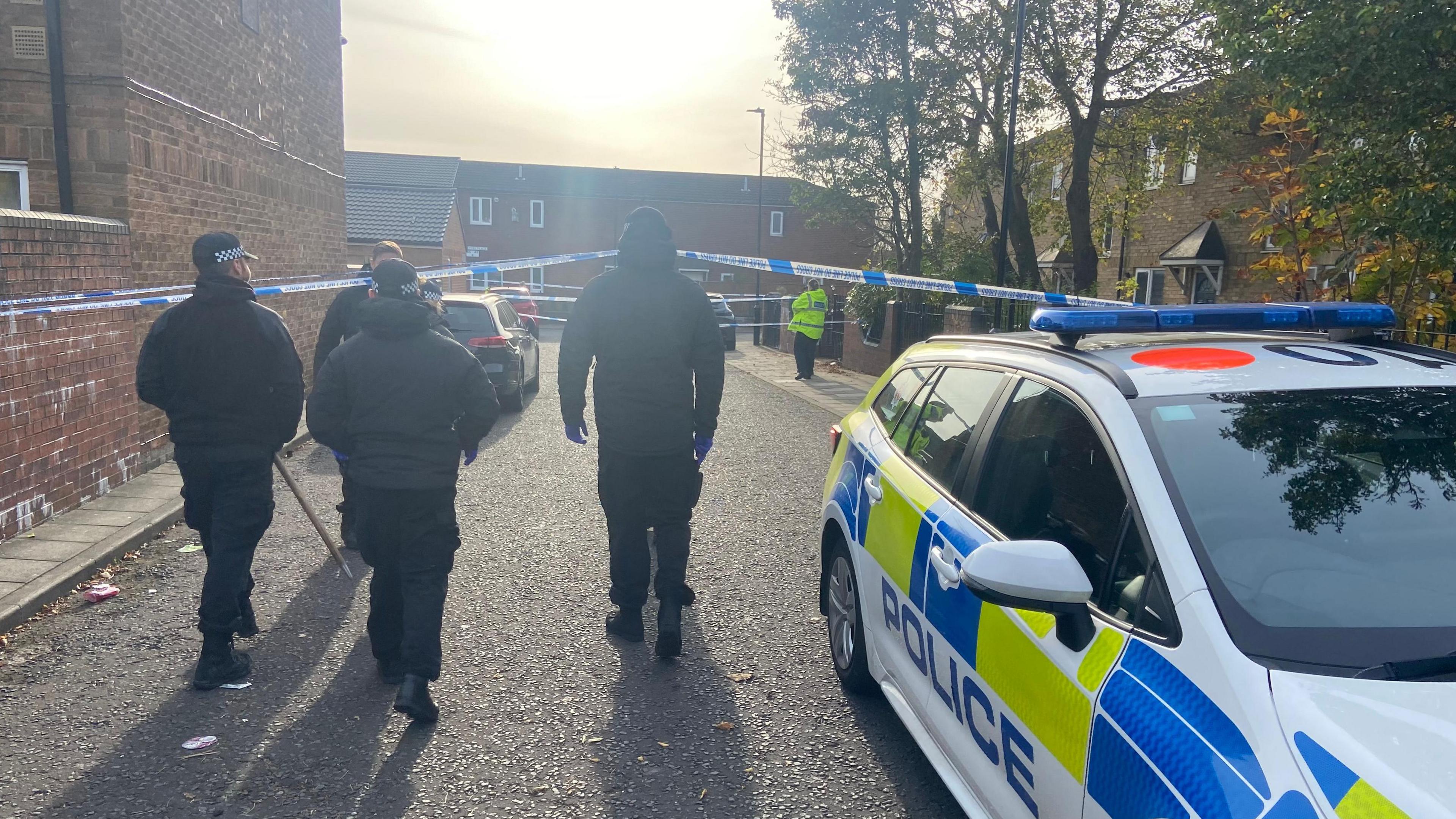 The back of four police officers wearing black, with a police car in the front right foreground. They are walking down a street with police tape stretched across it.