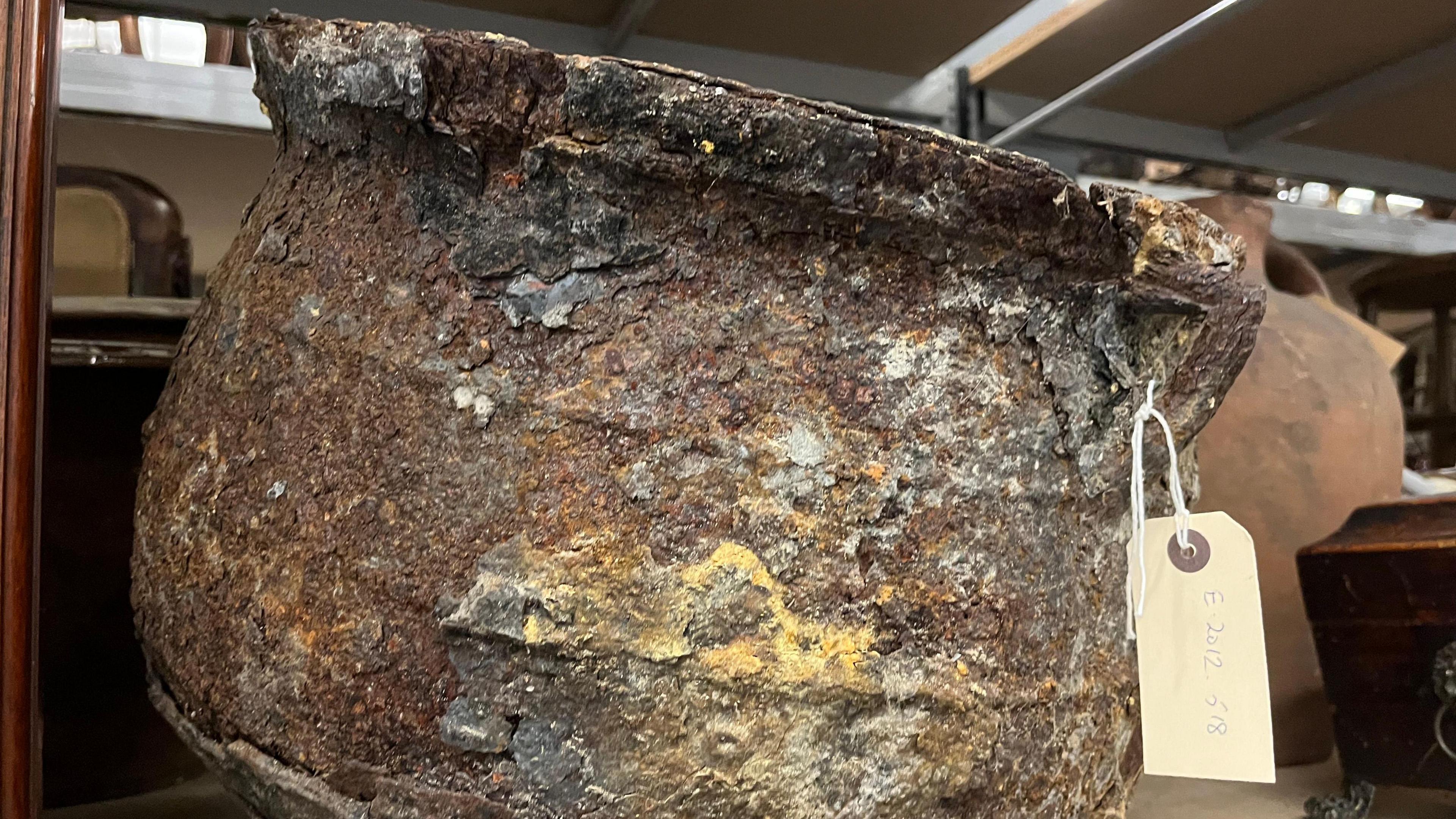 A heavily corroded, round metal pot with a wide opening and thick rim, showing extensive rust and flaking layers of oxidation in shades of brown, grey and yellow. The pot has a rough, uneven texture. A white tag with handwritten text is attached to the rim by a string, and the pot is placed on a shelf among other objects.