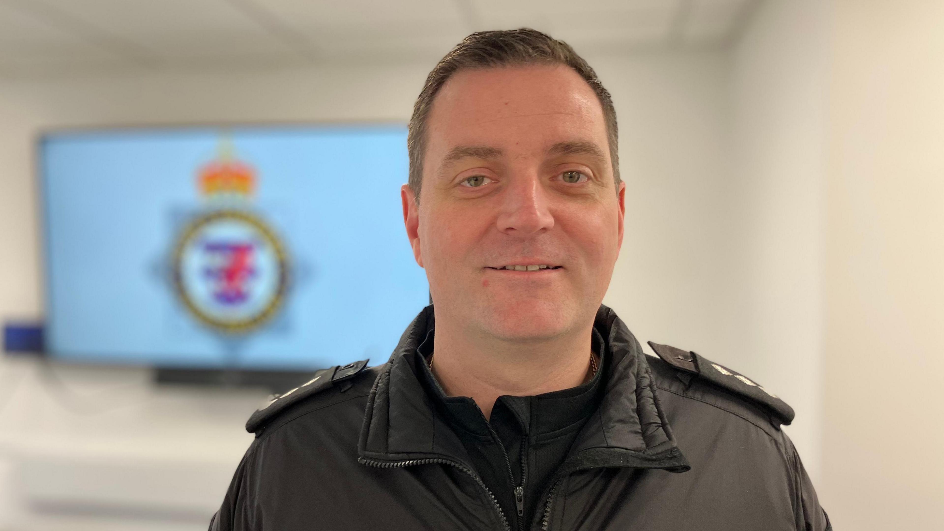 Ch Insp Rob Cheeseman wearing his black police uniform. He stands in front of an out-of-focus screen which has the Avon and Somerset Police logo on it.