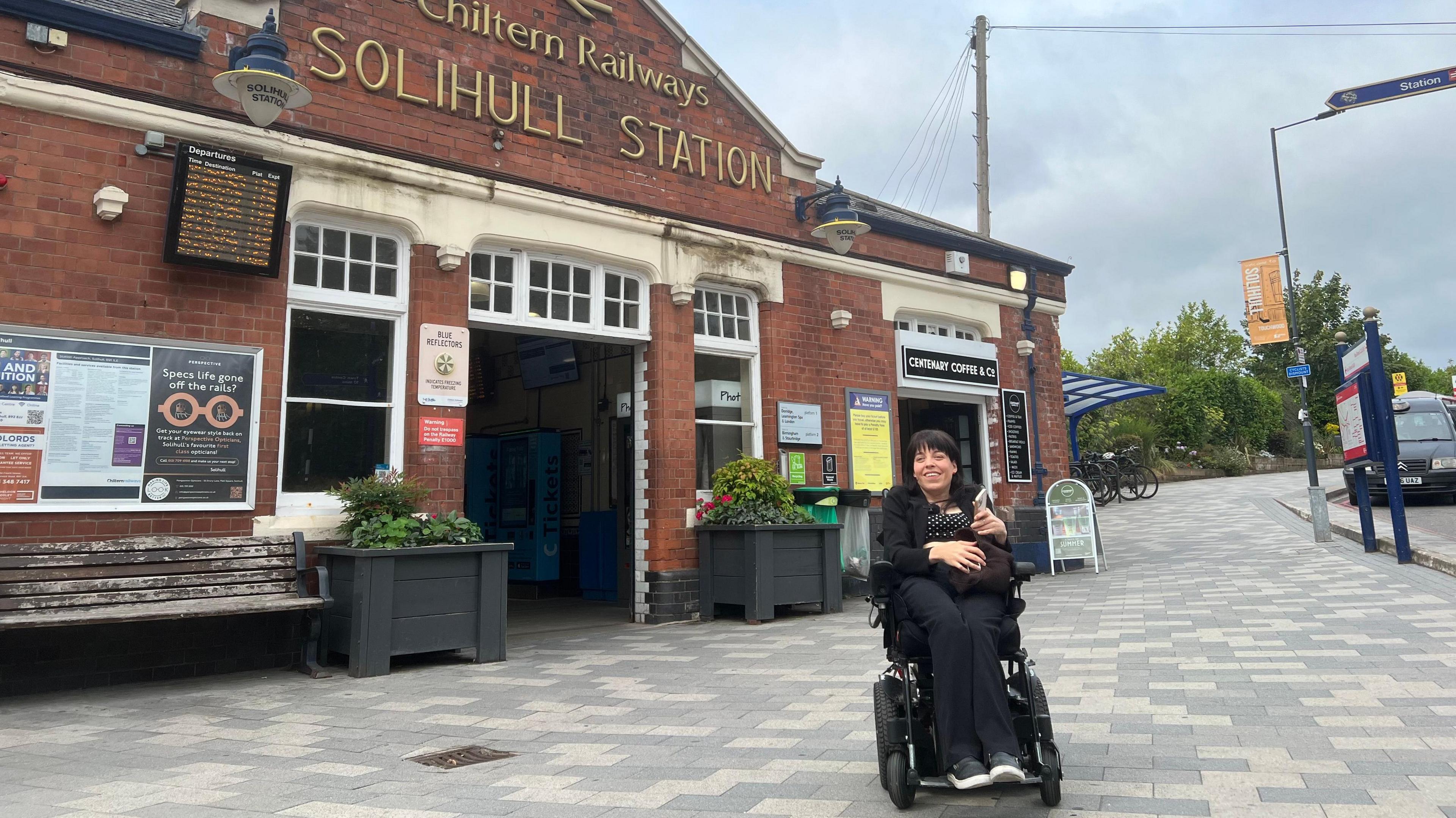 A woman in a wheel chair sitting infant of a railway station. She is wearing all black and has black hair. The station is red brick. 