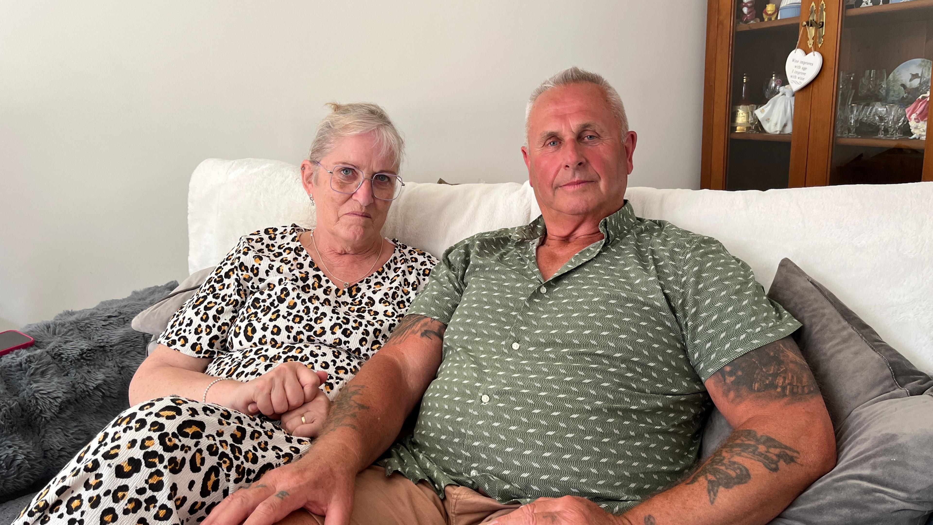 Nicki and Andy Duffield are sitting on a sofa looking at the camera. Nicki is wearing a white patterned dress and glasses. Andy is wearing a green and white patterned shirt.