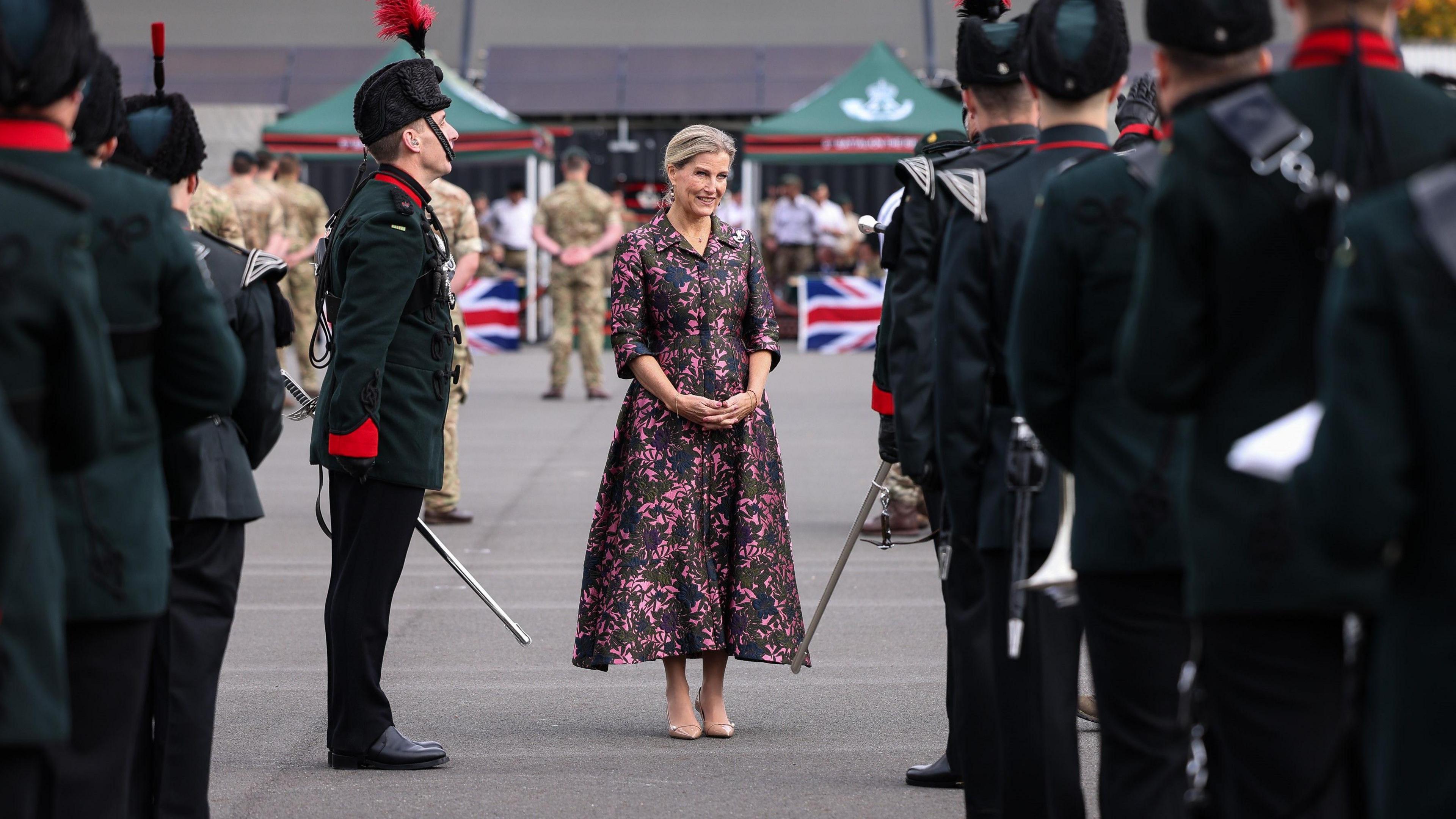 The Duchess of Edinburgh standing in a long embroidered dress in between soldiers in formal dress with swords