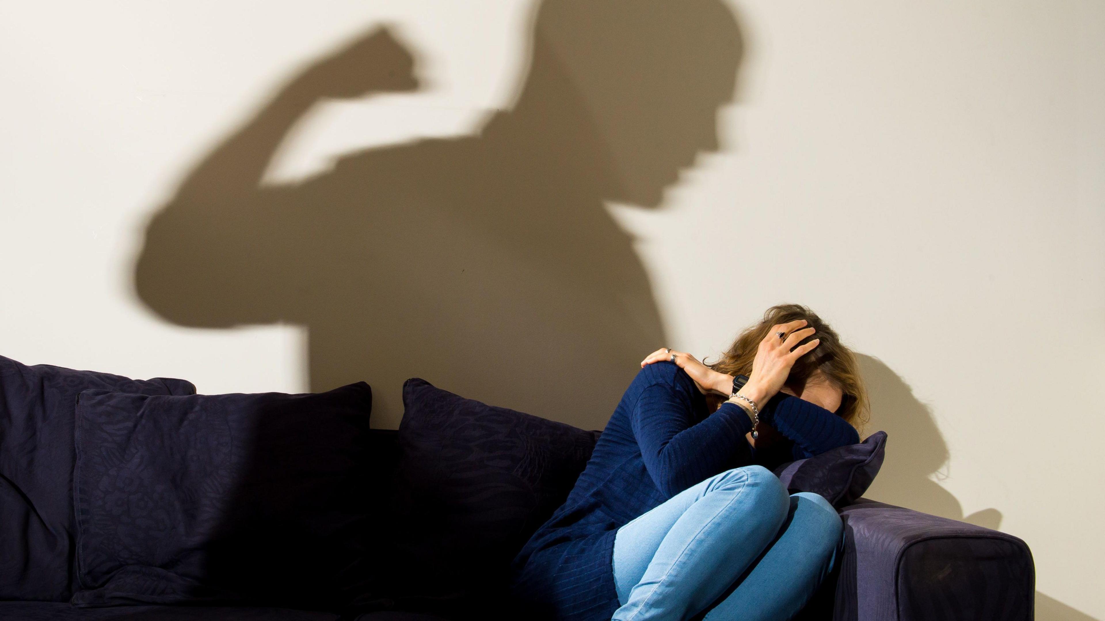 A silhouette of a man with a clenched fist seen over a woman cowering on the corner of a sofa.