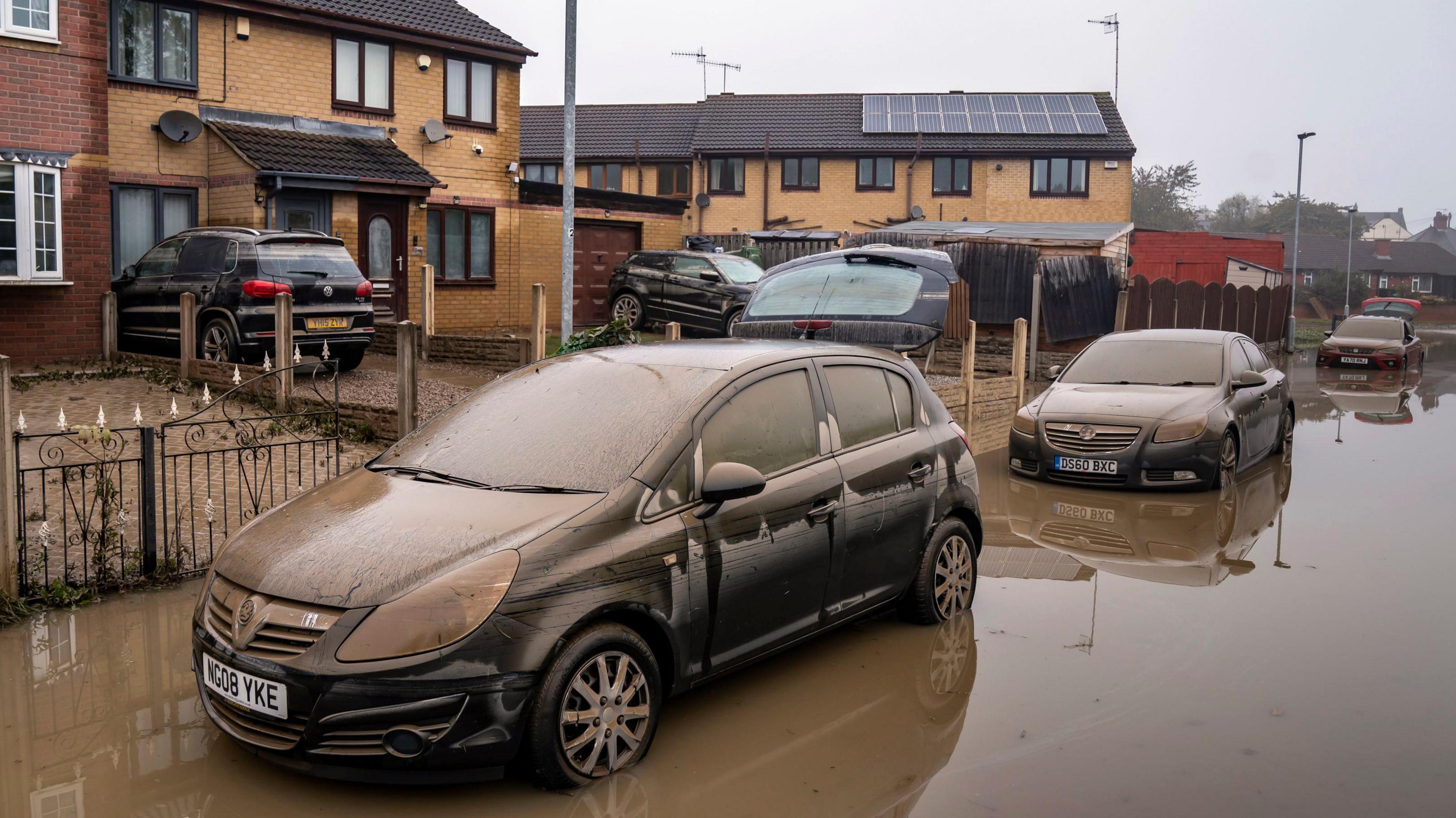 Cars covered in silt stand in floodwater on a street
