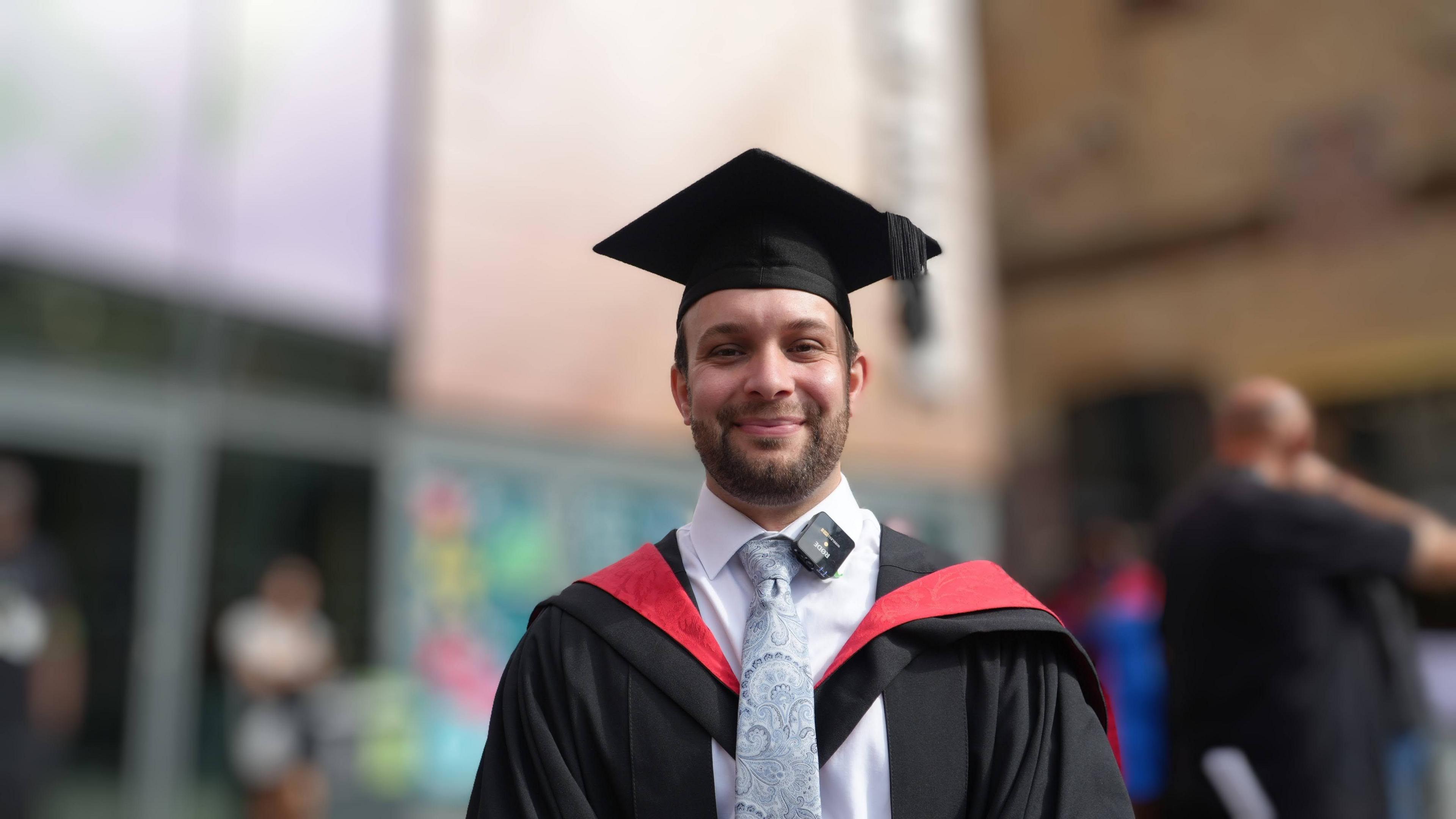 Michael Rose in a black graduation gown and hat, with red sash, and a grey paisley tie