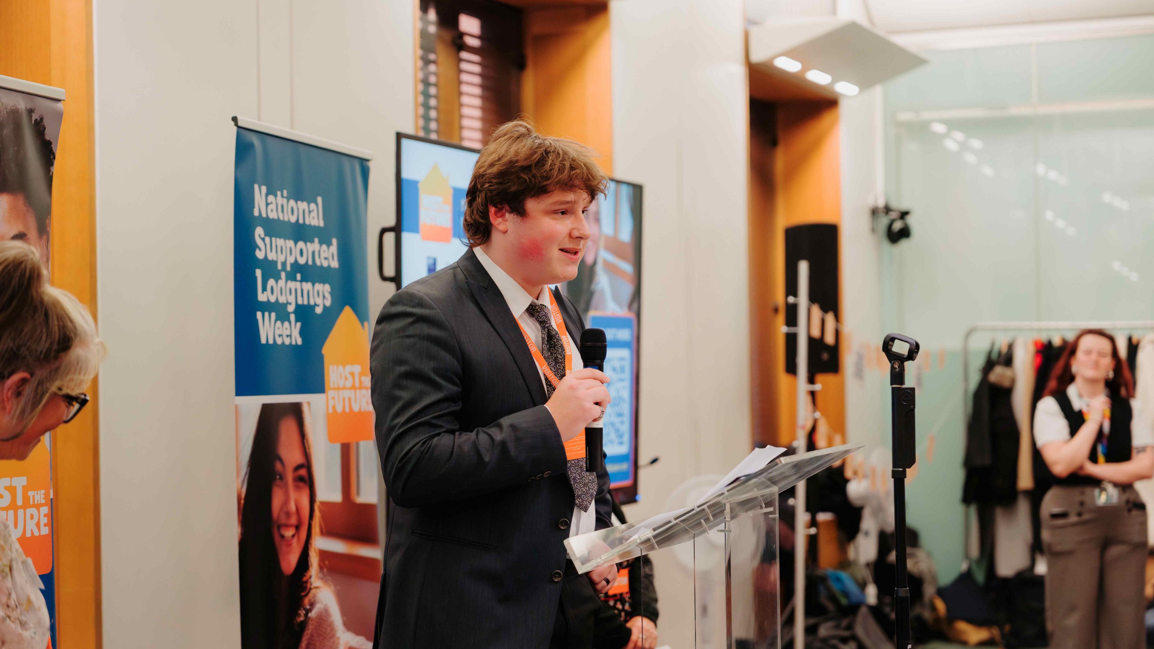 Young gentleman in a dark suit and tie holding a microphone. He's giving a speech in front of lots of people.