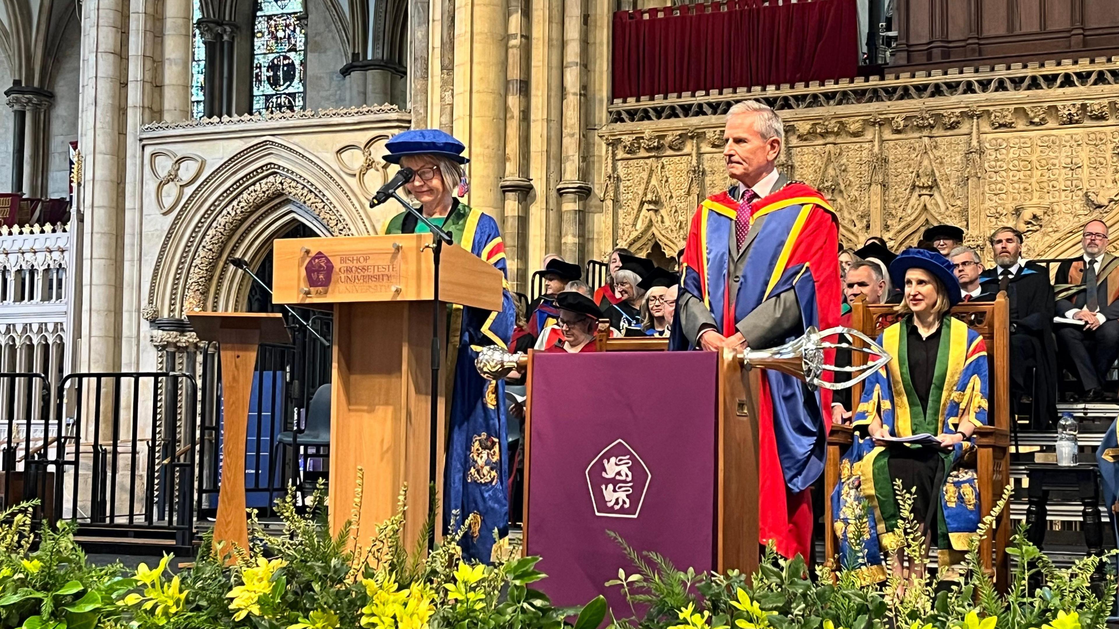 Peter Levy, wearing red, blue and yellow graduation robes, prepares to receive his doctorate in front of a grand, stone doorway at Lincoln Cathedral. Behind him sit figures from ishop Grosseteste University, also dressed in colourful robes.