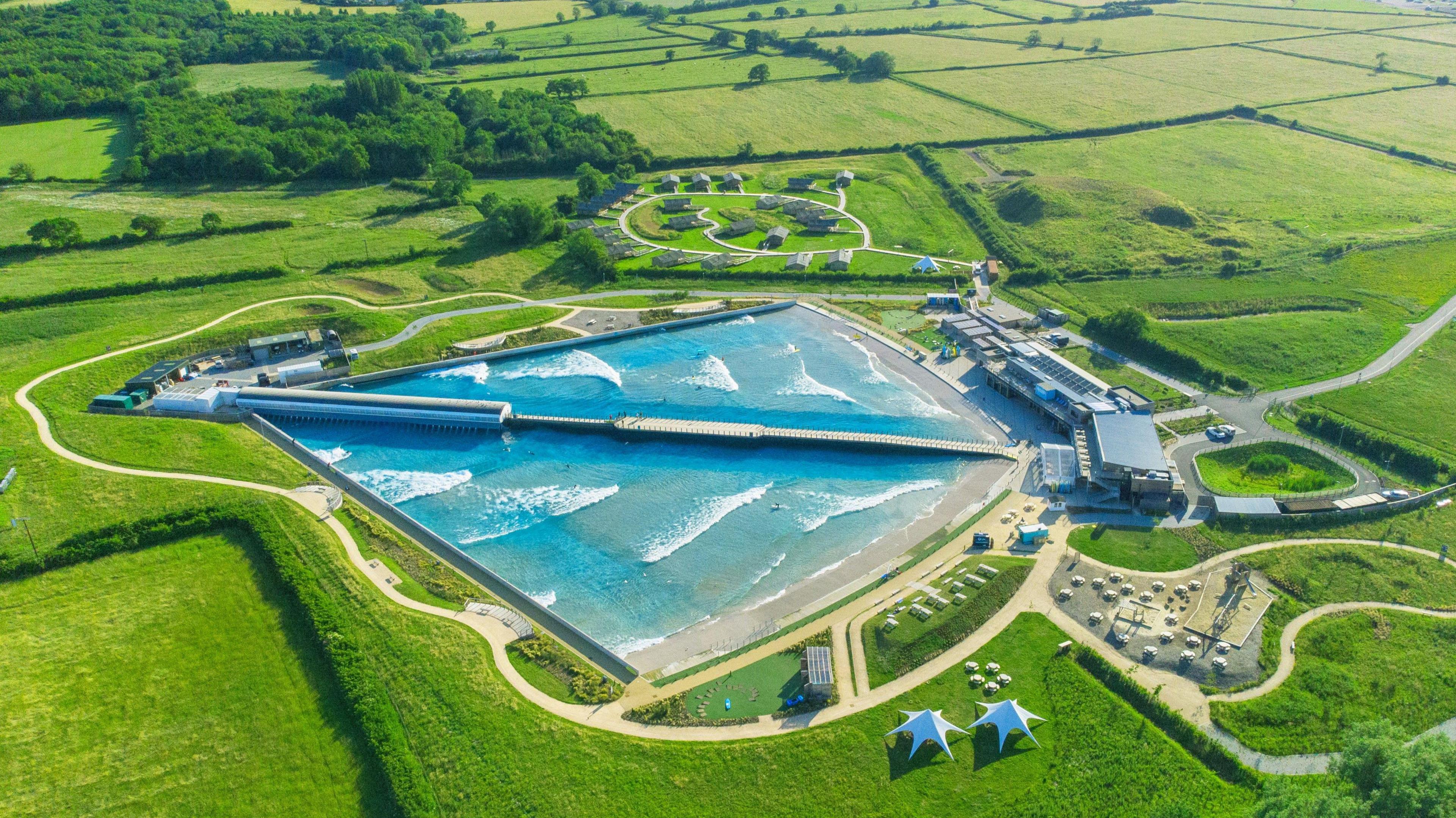 An aerial view of The Wave. It is a kite-shaped lake in the middle of lots of bright green fields. The water is bright blue and there is a wooden pier running down the middle. At the base of the diamond there is a clubhouse and café, along with several tents and outbuildings. 