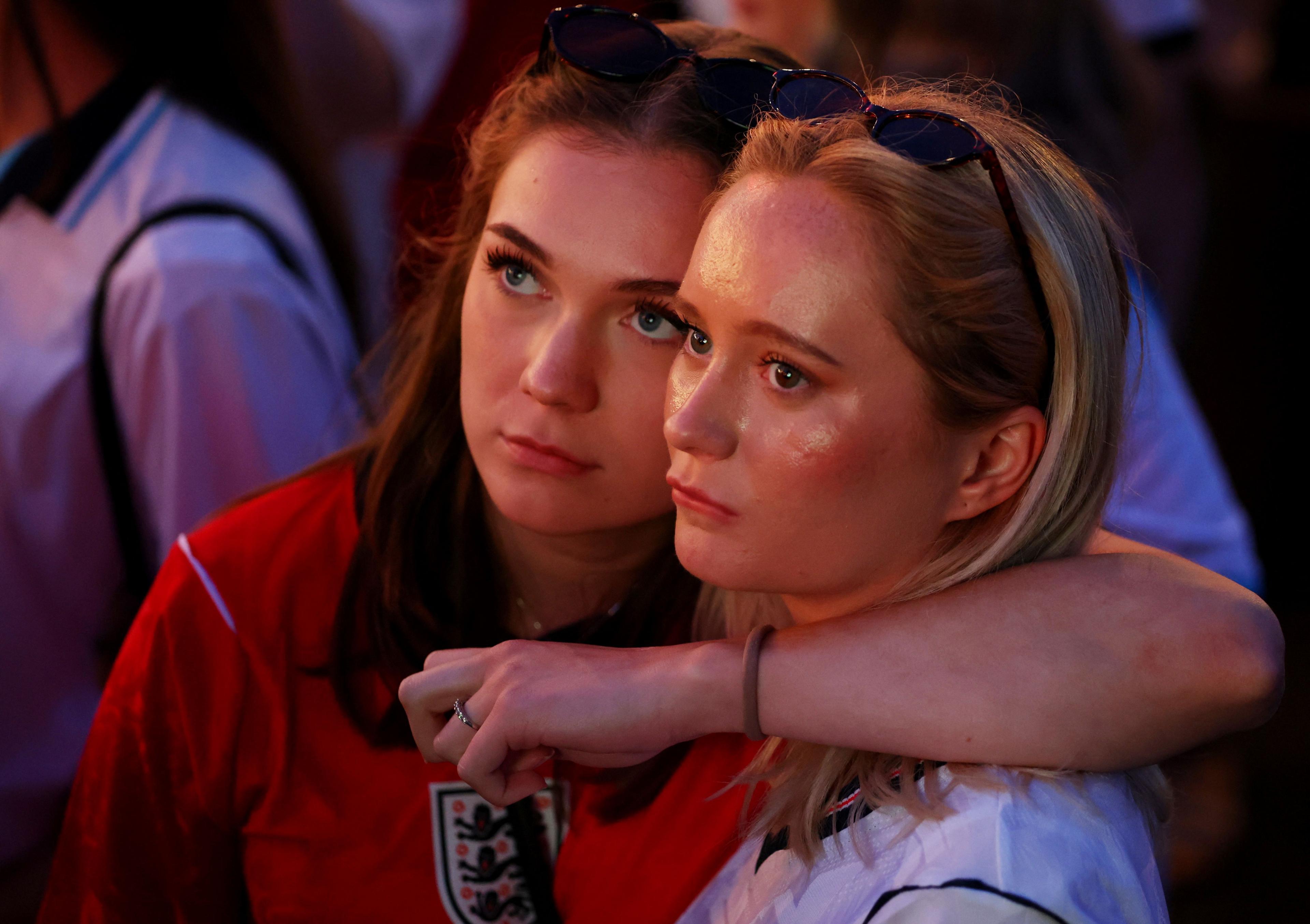Two England fans look dejected after the match. One woman in a red England shirt  with sunglasses pinned on top of her head has her arm wrapped around another woman in a white top.