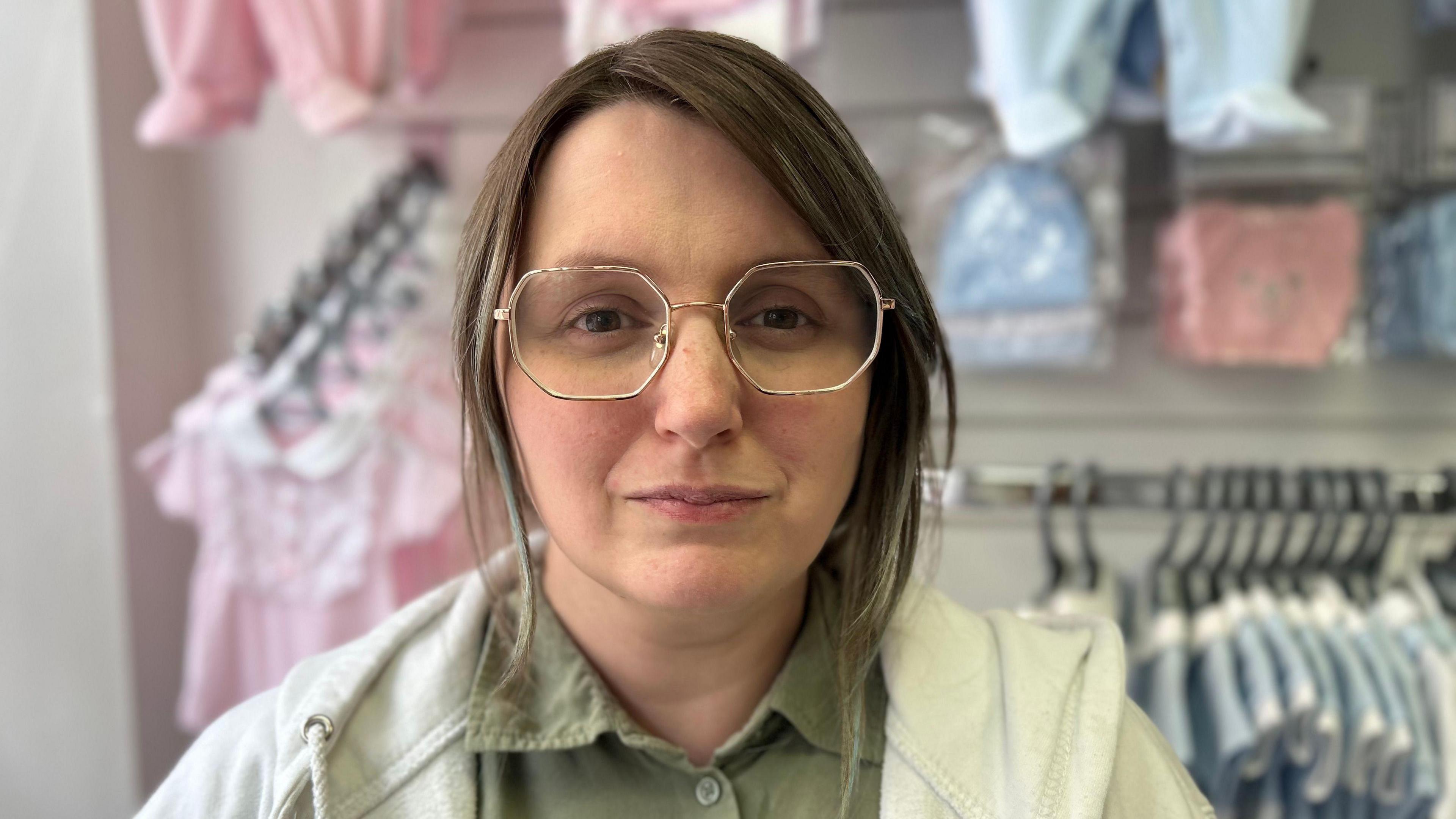 A girl wearing glasses with her hair besides her ears wearing a green shirt and a white hoodie, stood behind baby clothes sold in the shops.