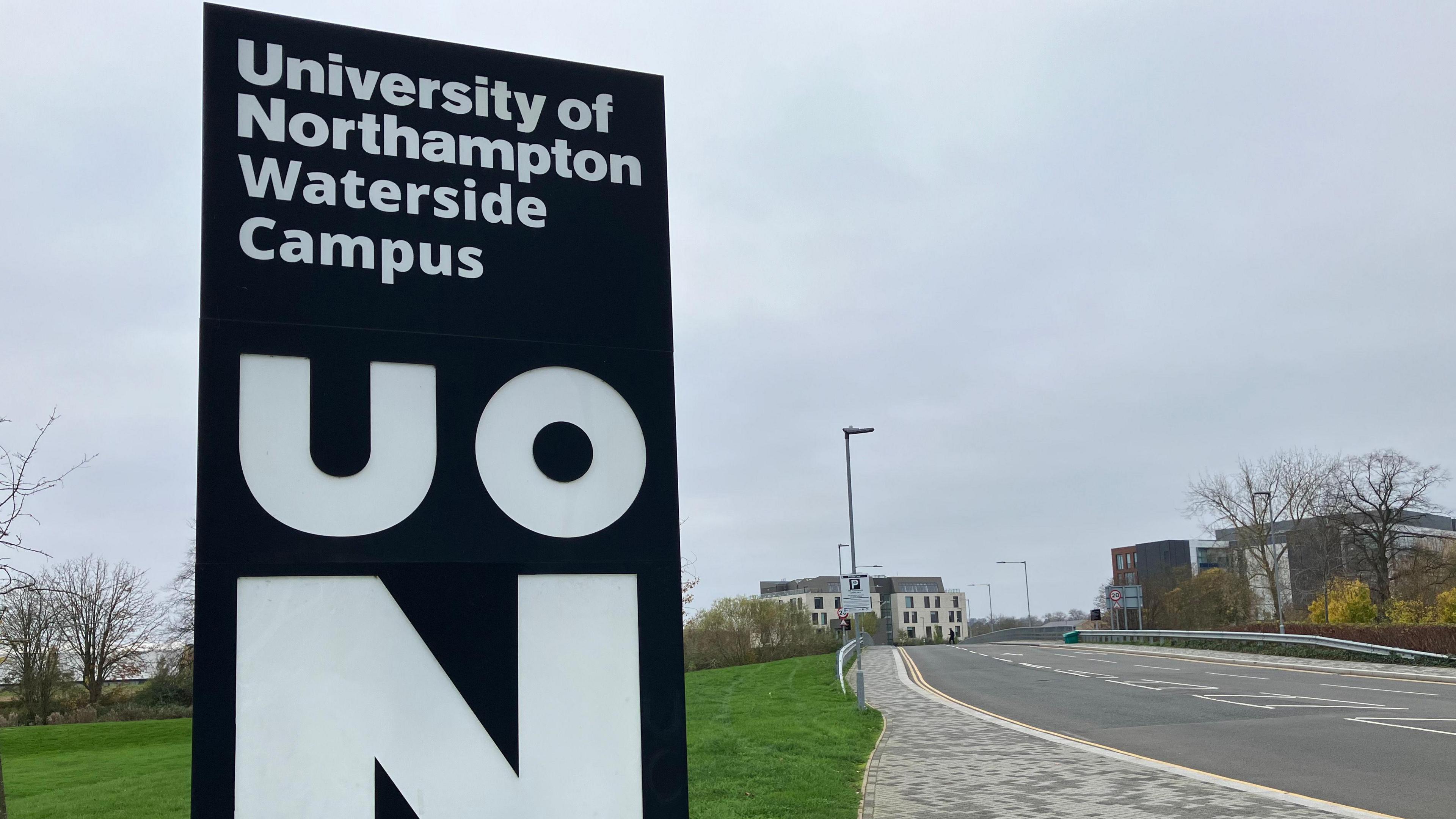 A black sign for the University of Northampton by a road.