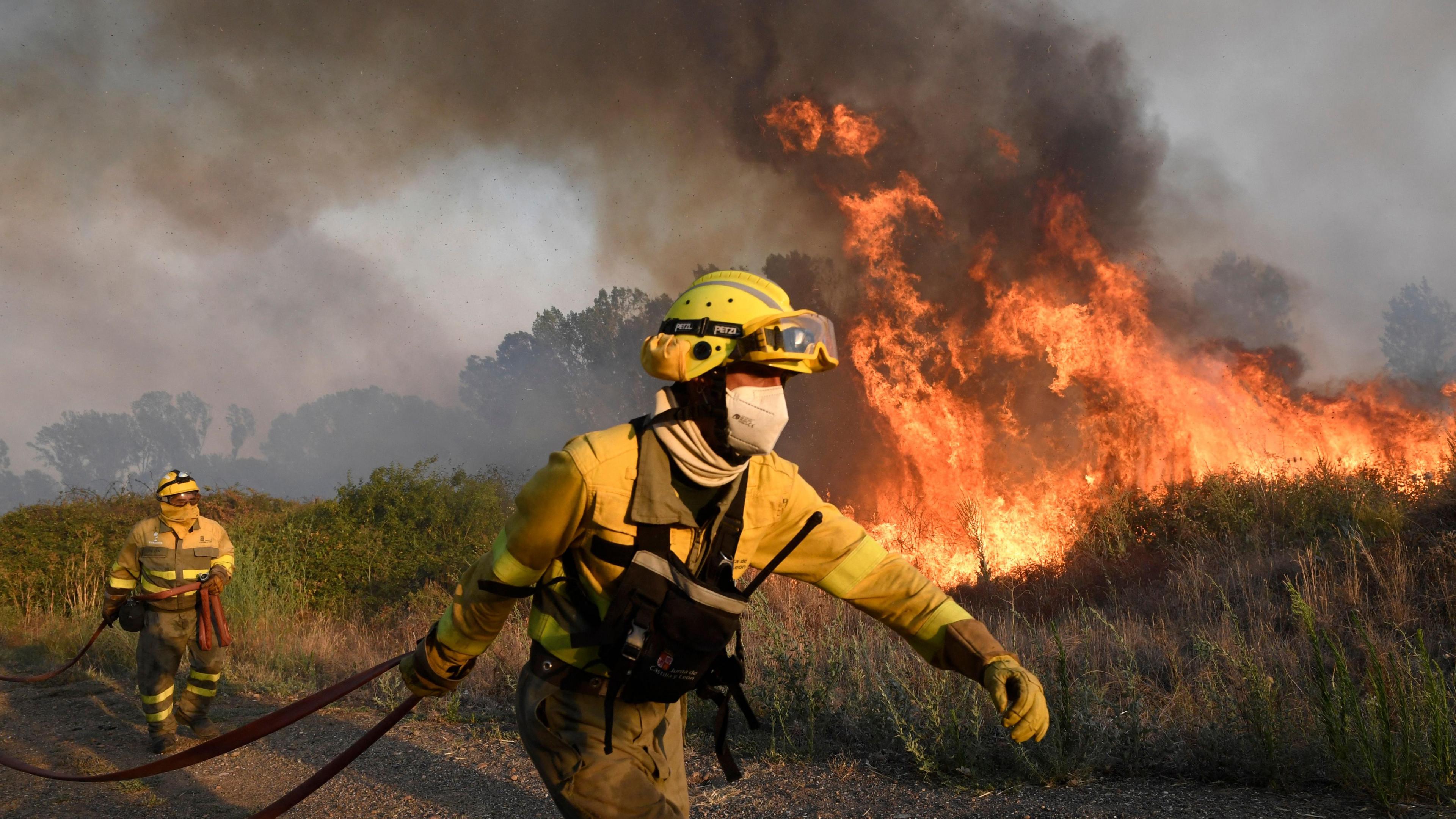 Two firefighters wearing protective clothing and headgear are carrying dark red firehoses near a village in northern Spain. They are walking along a track beside a grassy area which is on fire. 