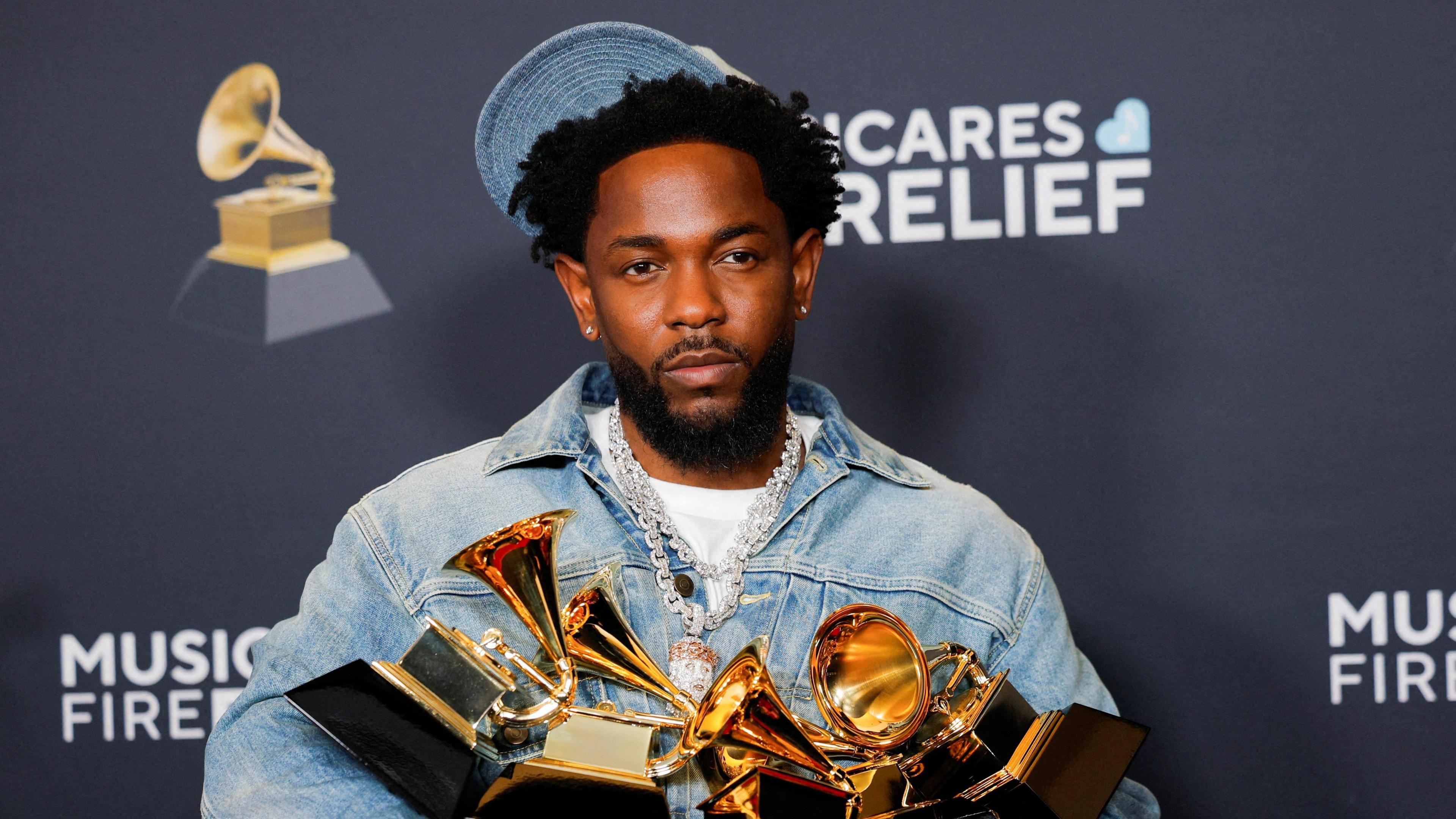 Kendrick Lamar poses for a picture while carrying his five Grammy awards. He is wearing a denim jacket and cap with a bejeweled silver chain around his neck.