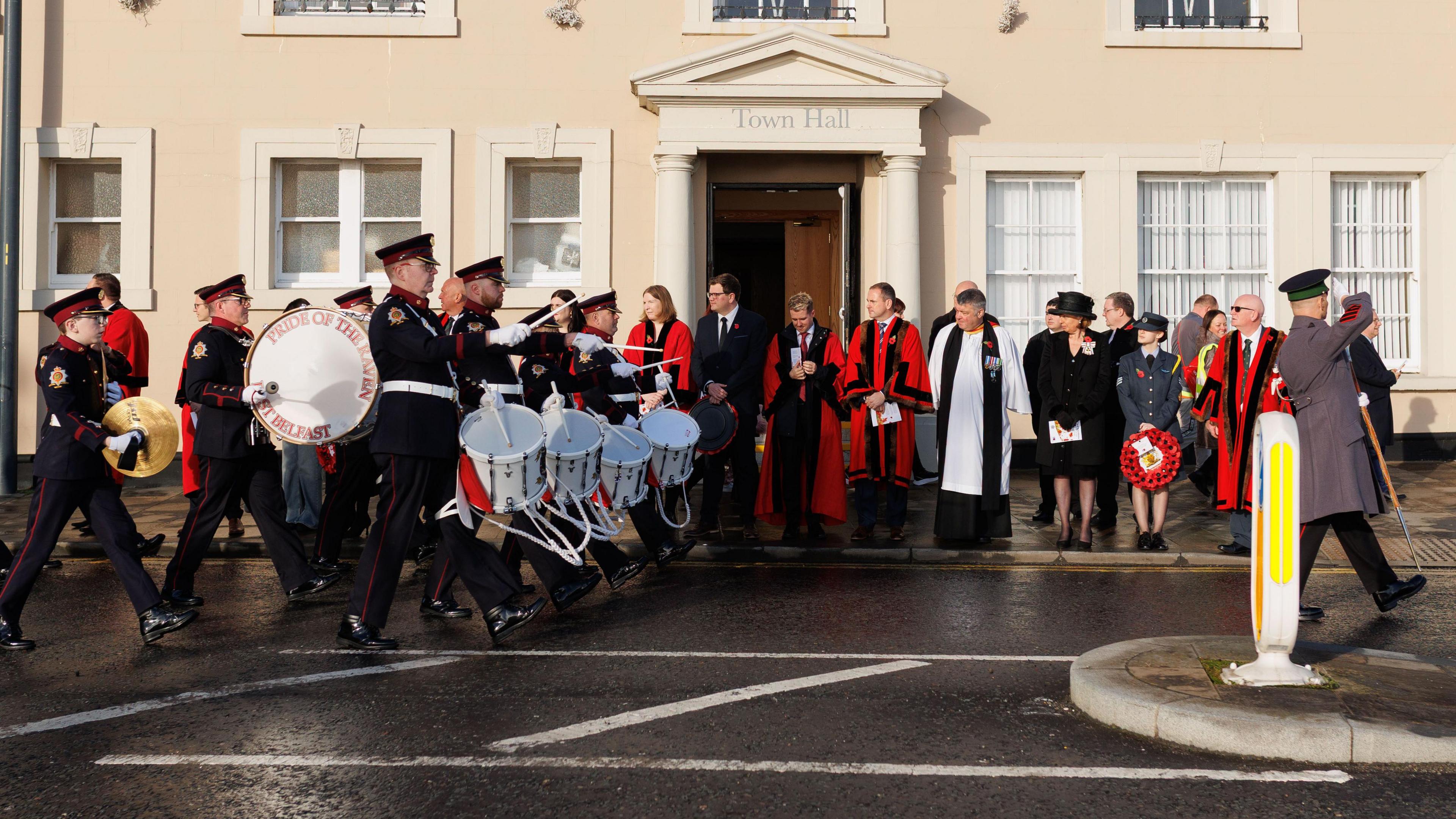 A parade is walking through a road. The people in front are holding instruments. and wearing black jackets with back trousers. ome people are holding poppy wreaths. 