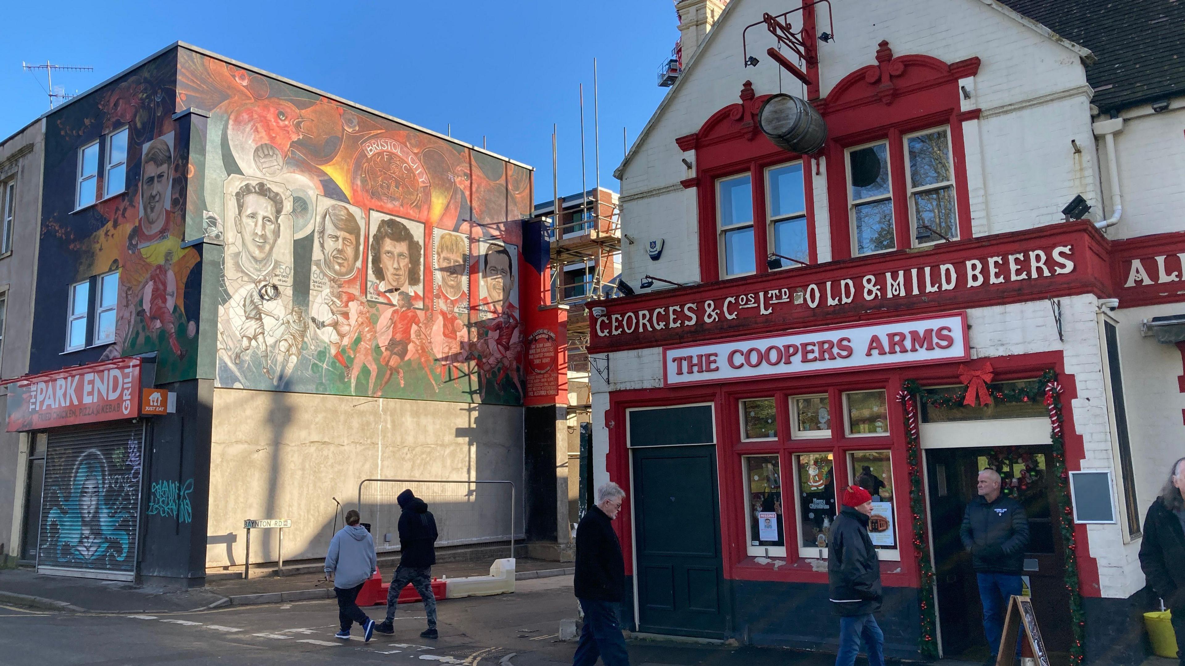 The mural, pictured from across the street and next to The Coopers Arms.  The pub has a red and white facade and there are people in coats and hats walking past. The sky is blue and the mural is visible on the side of a two-storey building. 