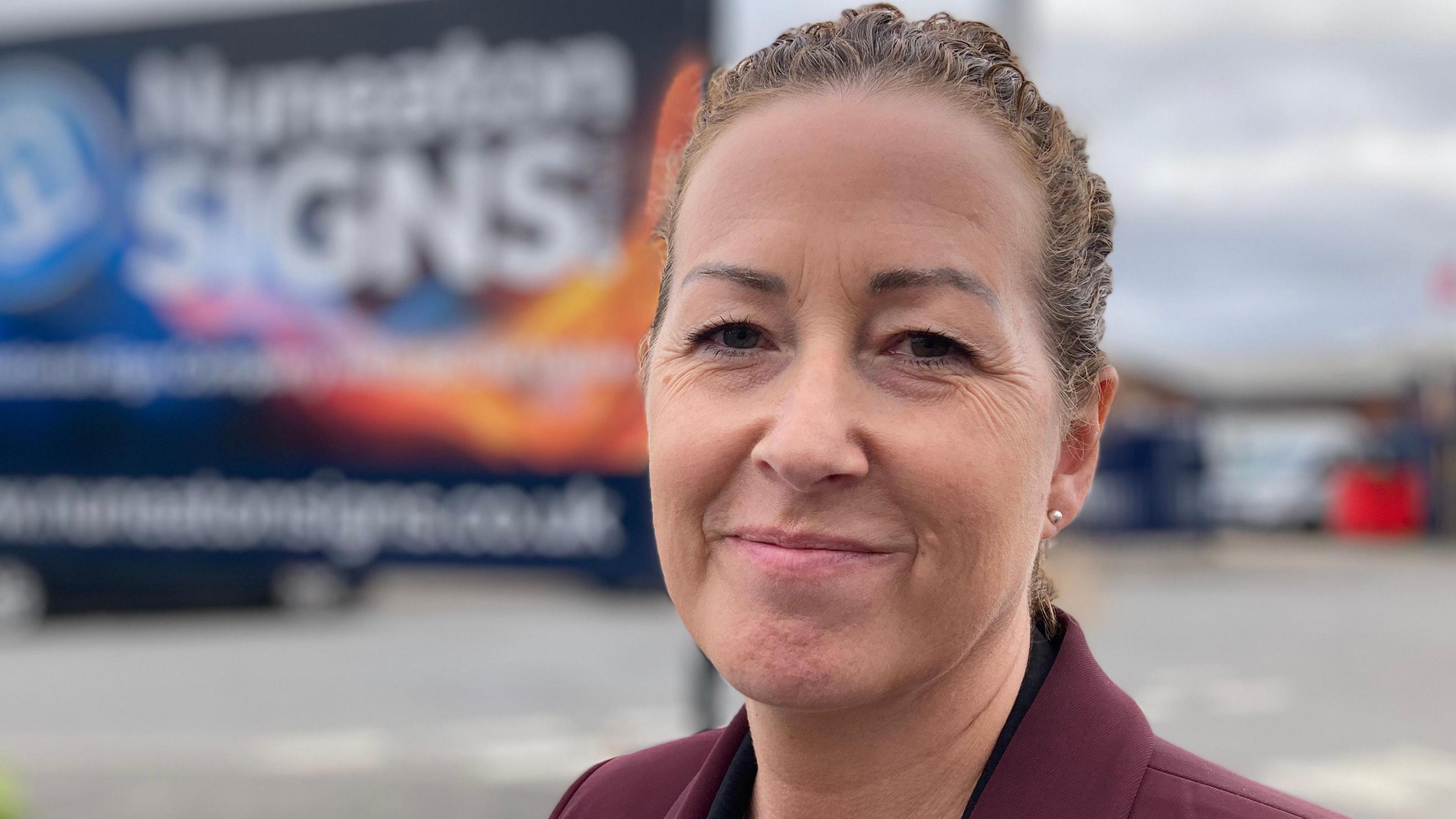 A woman smiles toward the camera , she is wearing a burgundy jacked and black blouse, behind her is a sign with Nuneaton Signs written on it in large text.