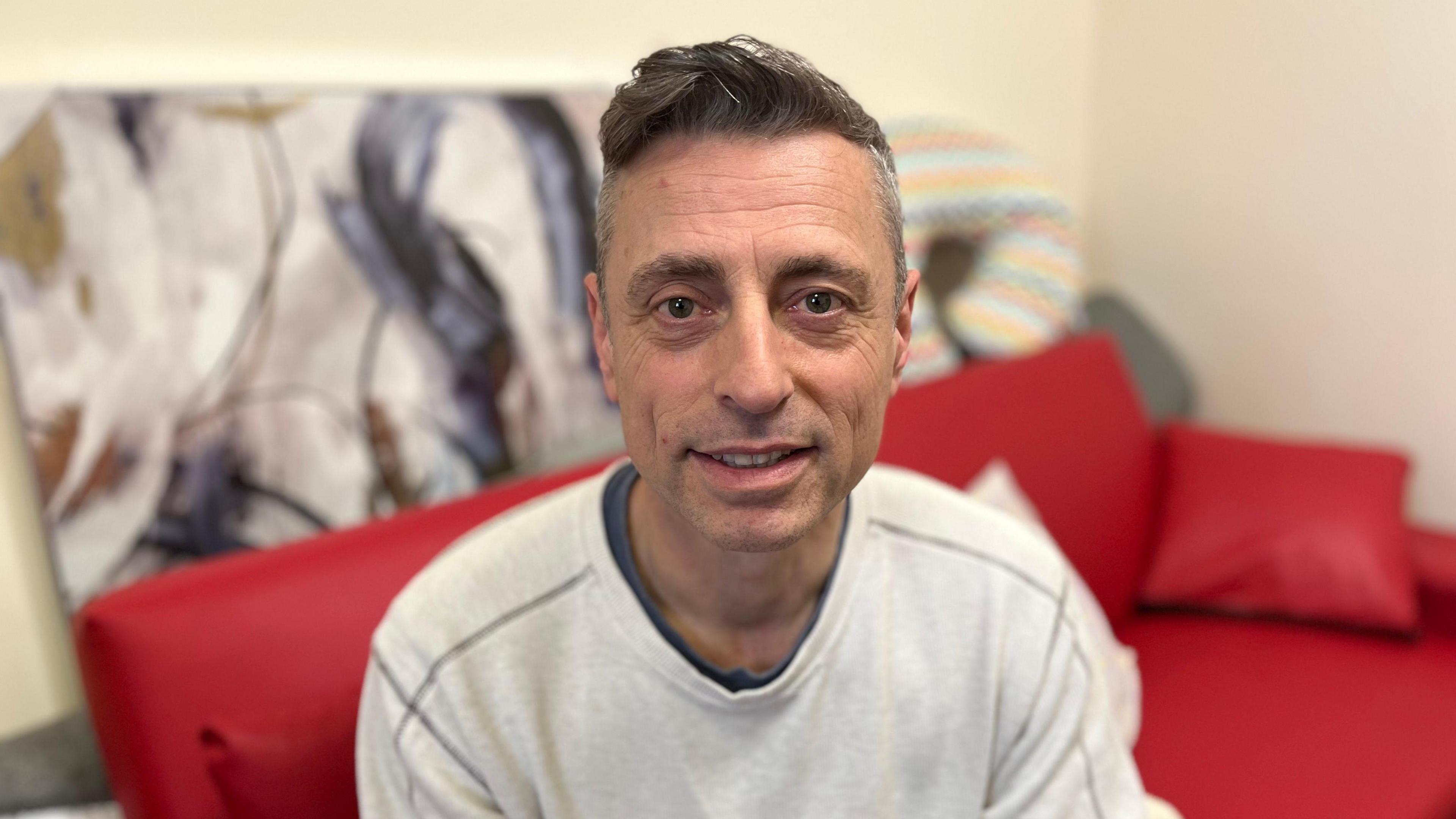 Mark Bowering smiles at the camera as he sits on a red sofa. He is wearing a light grey jumper and there is a painting leaning against a wall behind the sofa. 