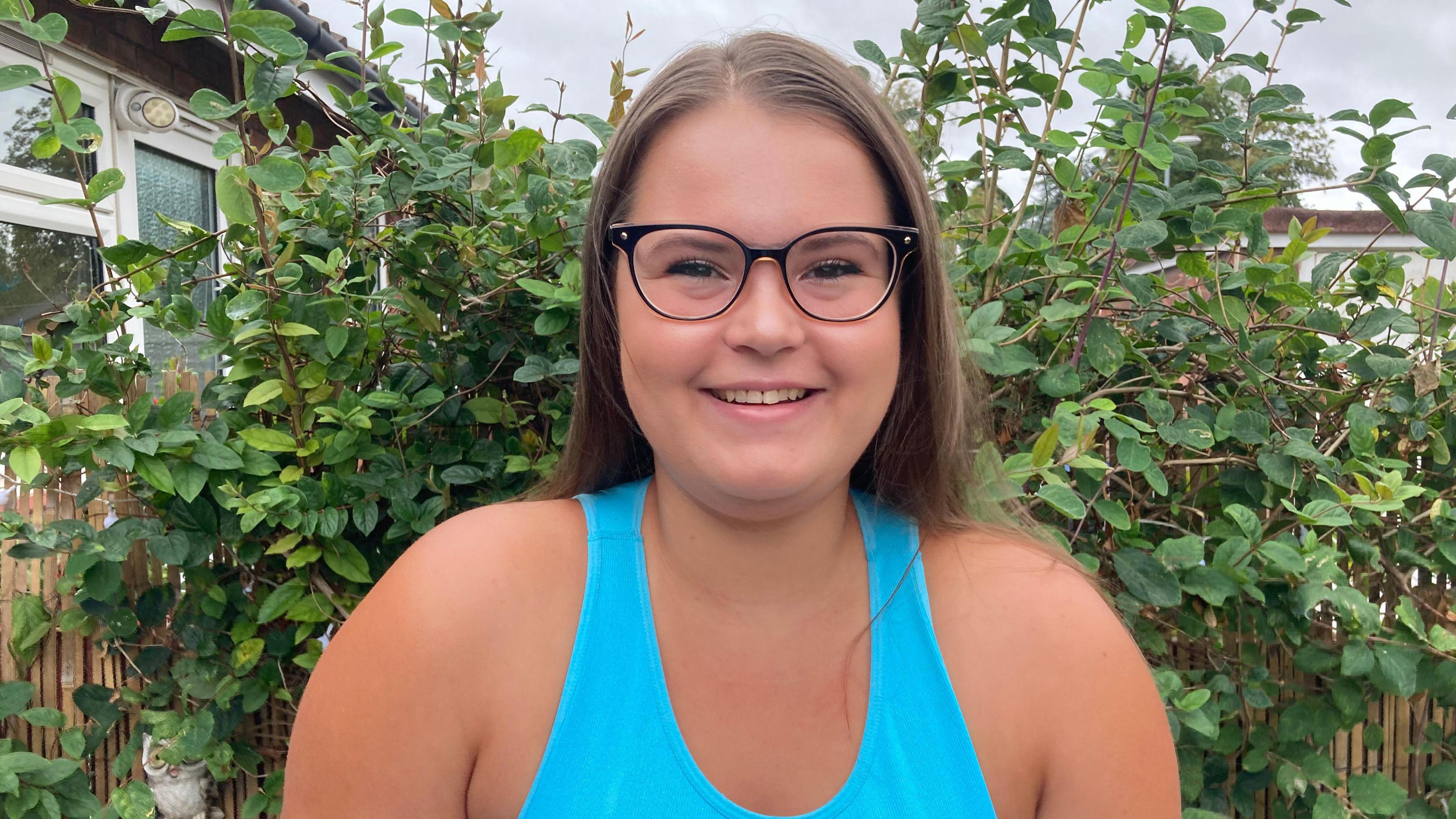 Jordan Till smiling at the camera while stood outside. She is wearing a bright blue vest top, black wide framed circle glasses, and has long light brown hair that has been pushed behind her shoulders.