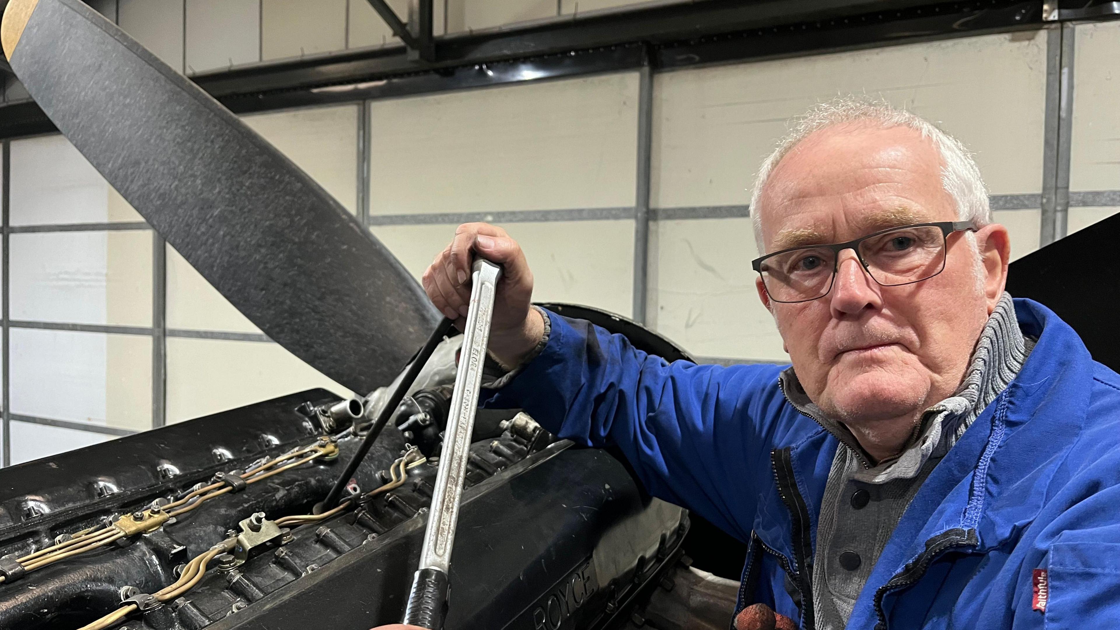 Volunteer Rob stood next to one of the aircraft's engines holding a tool. He is looking at the camera, has short grey hair and black glasses. He is wearing a grey sweater and a blue work jacket. 