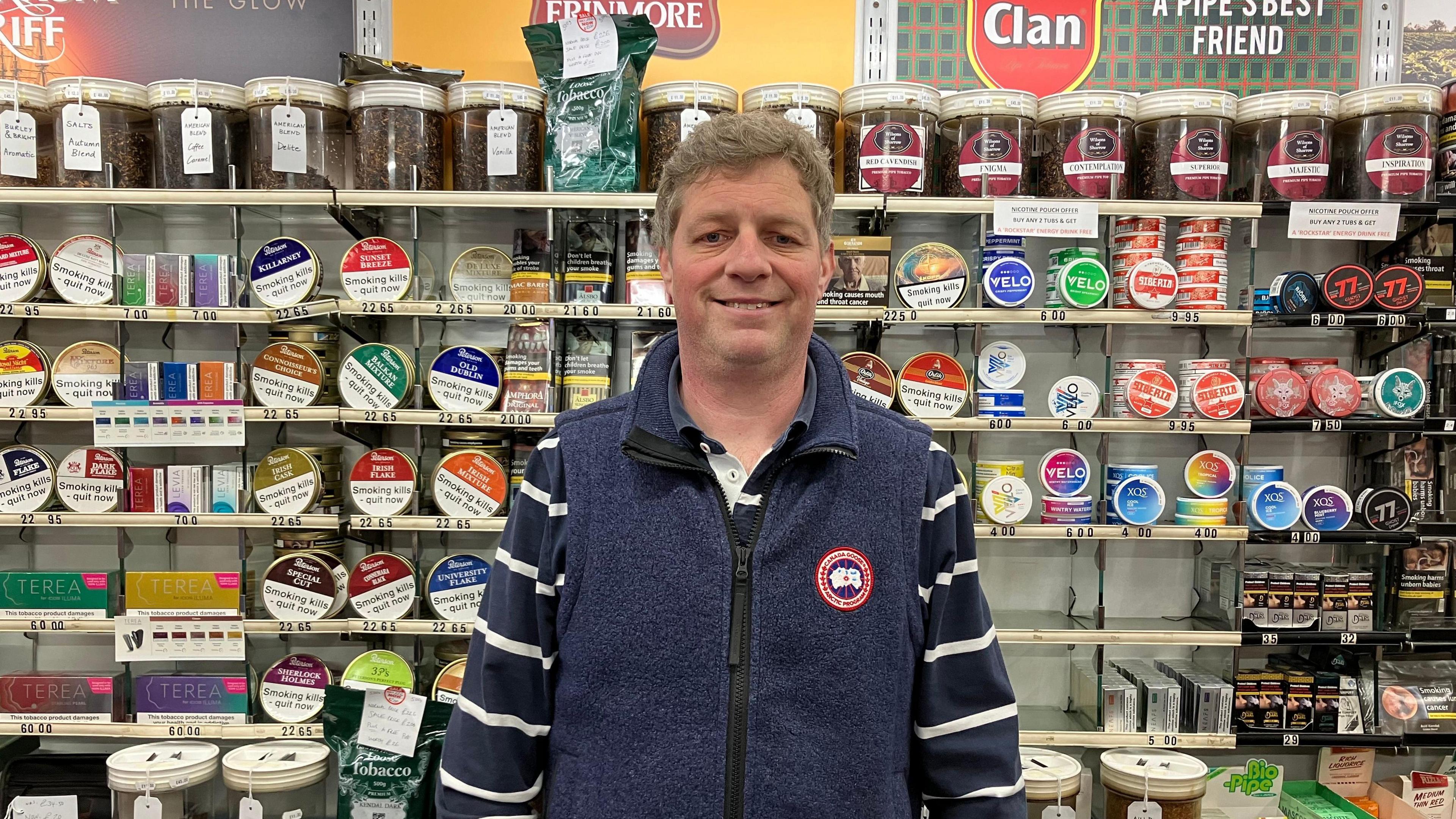 A man with brown and grey hair in a blue gilet and navy and white striped top standing behind a shop counter. Behind him tins of tobacco and other smoking-related paraphernalia line shelves. 