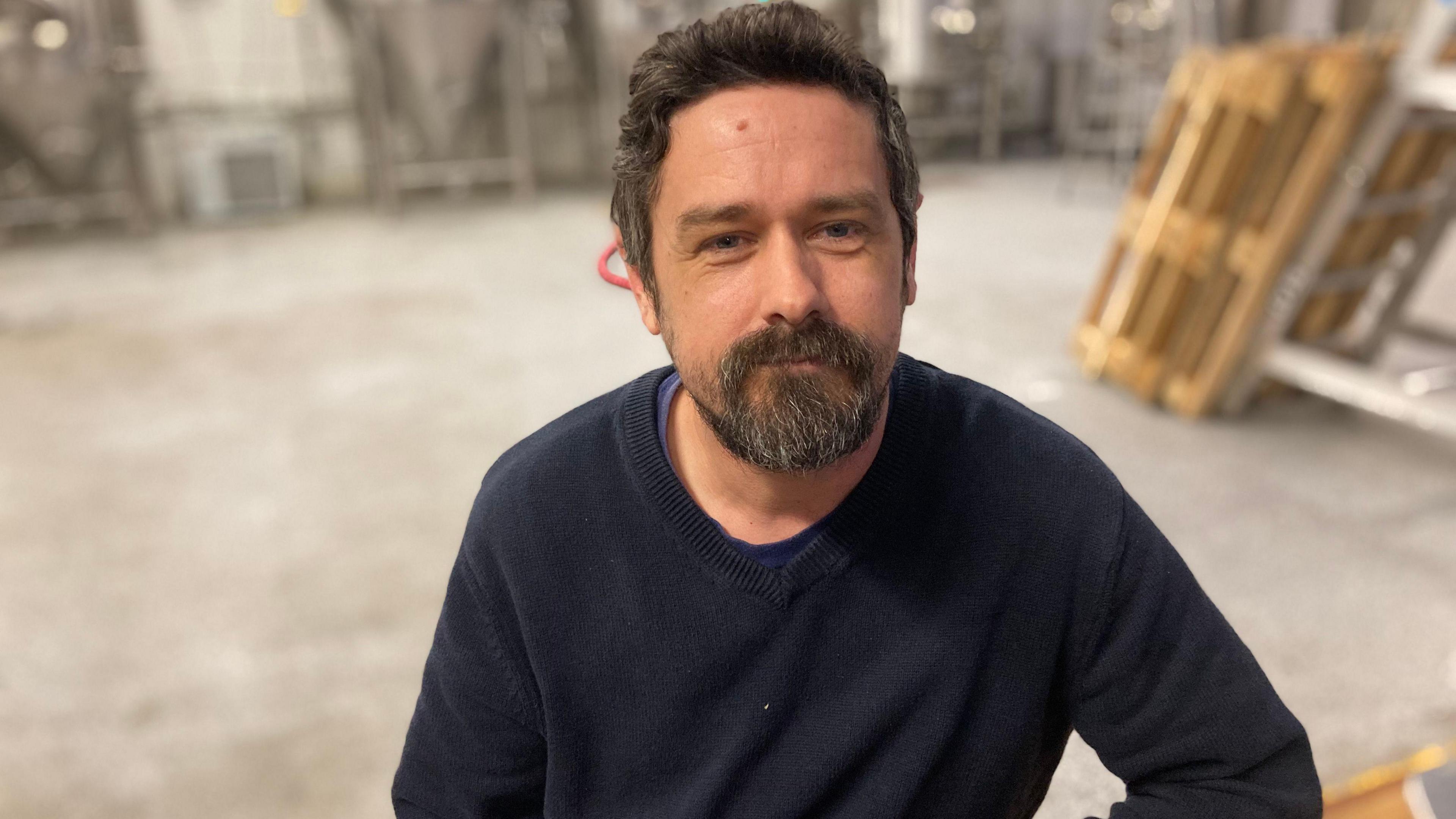 A man with brown hair and a beard weaering a blue jumper looks into the camera.