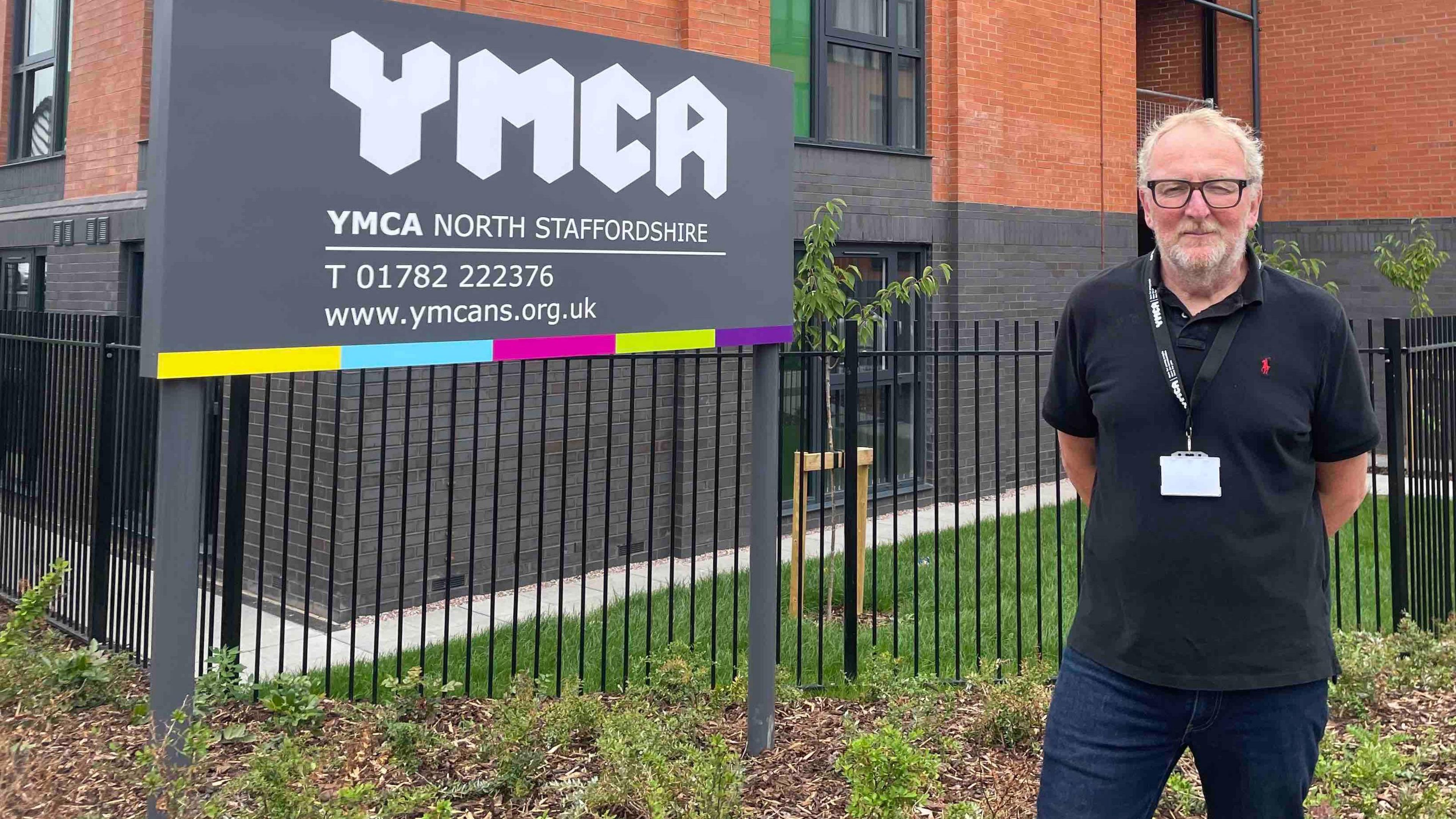 Daniel Flynn, a man with receding white hair and wearing a black polo shirt and dark blue jeans, is stood outside a block with windows and different floors, next to a grey sign which has white lettering that reads YMCA North Staffordshire.