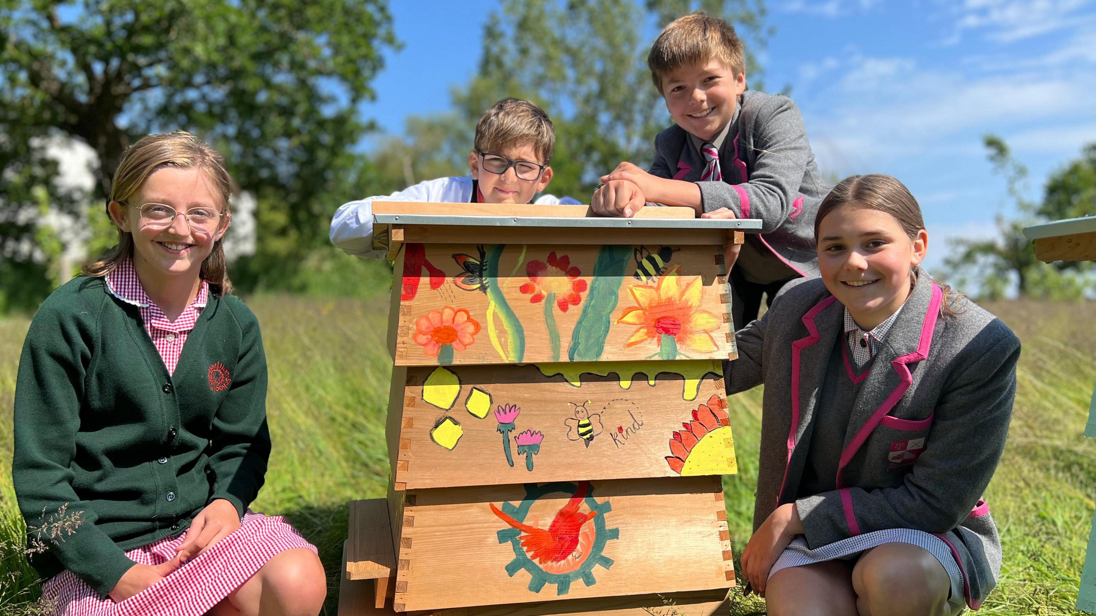 Eira and Esme kneeling next to a hand-painted bee hive, with Joao and Henry leaning on top