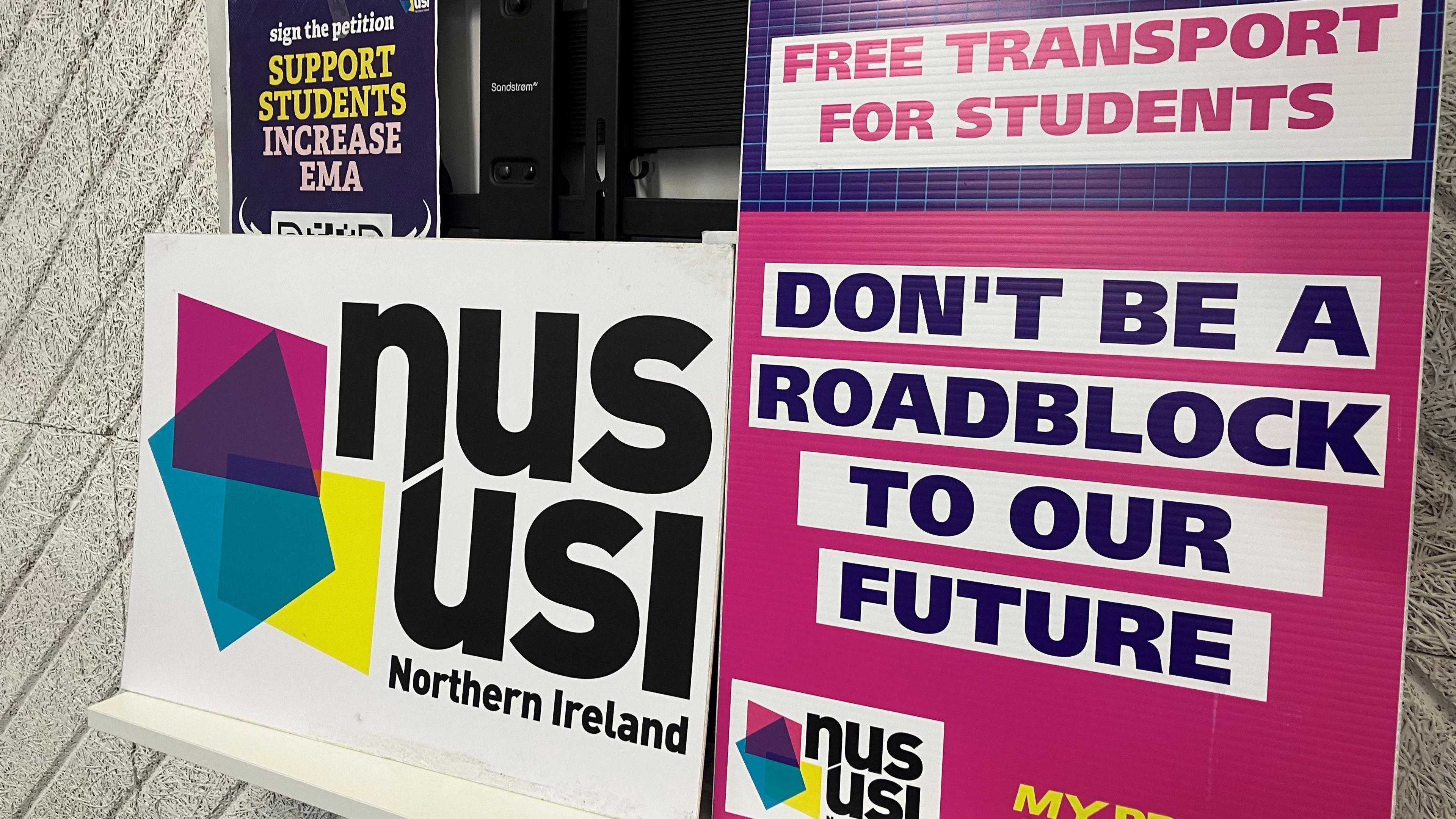 NUS-USI placards leaning against a wall. They are coloured - white, pink, yellow and blue. One says NUS-USI Northern Ireland, another says 'Don't be a roadblock to our students'.