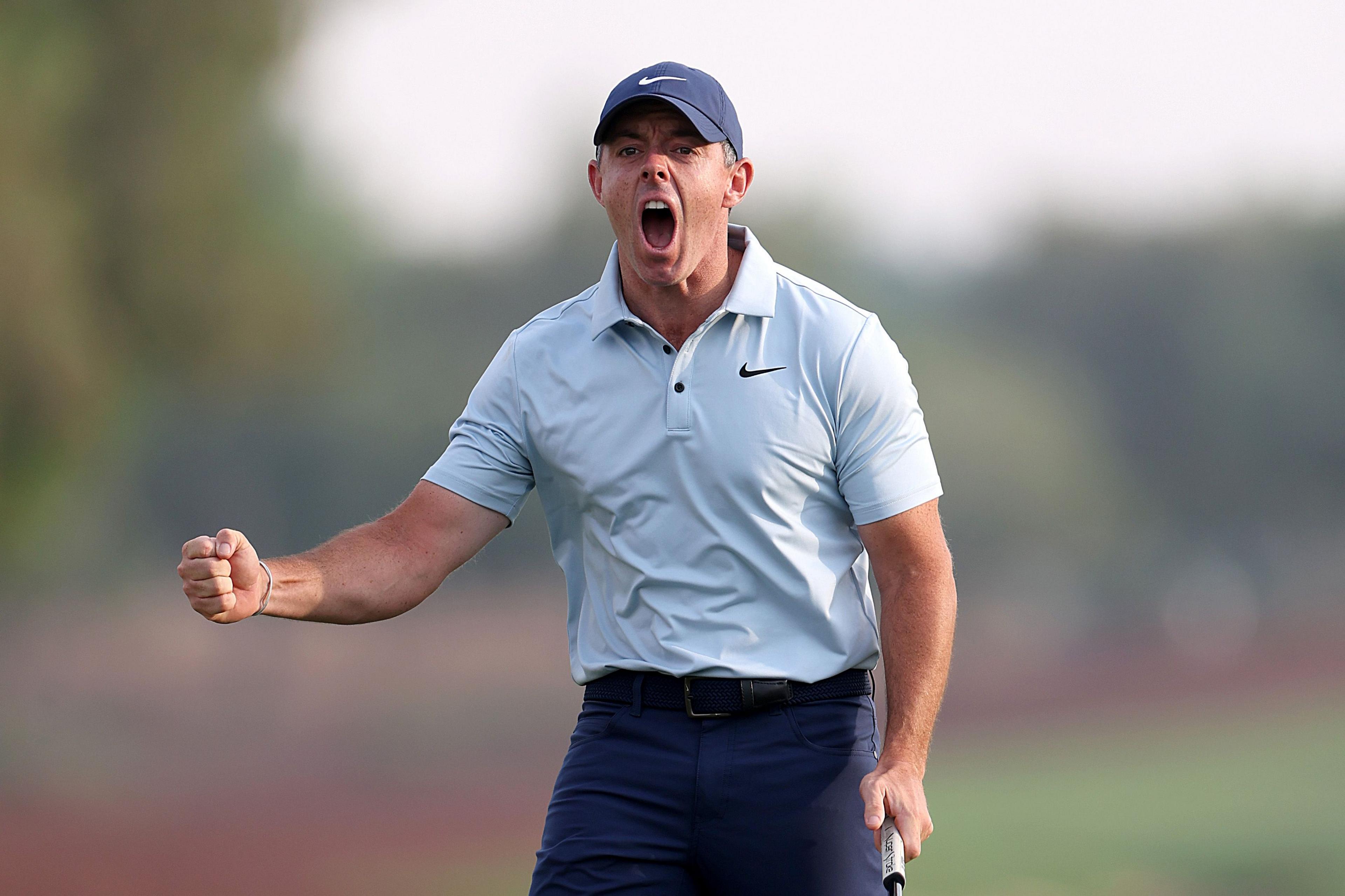 Rory McIlroy wearing a light blue collared shirt and dark pants, holding a golf club and clenching a fist in a celebratory gesture on a golf course.