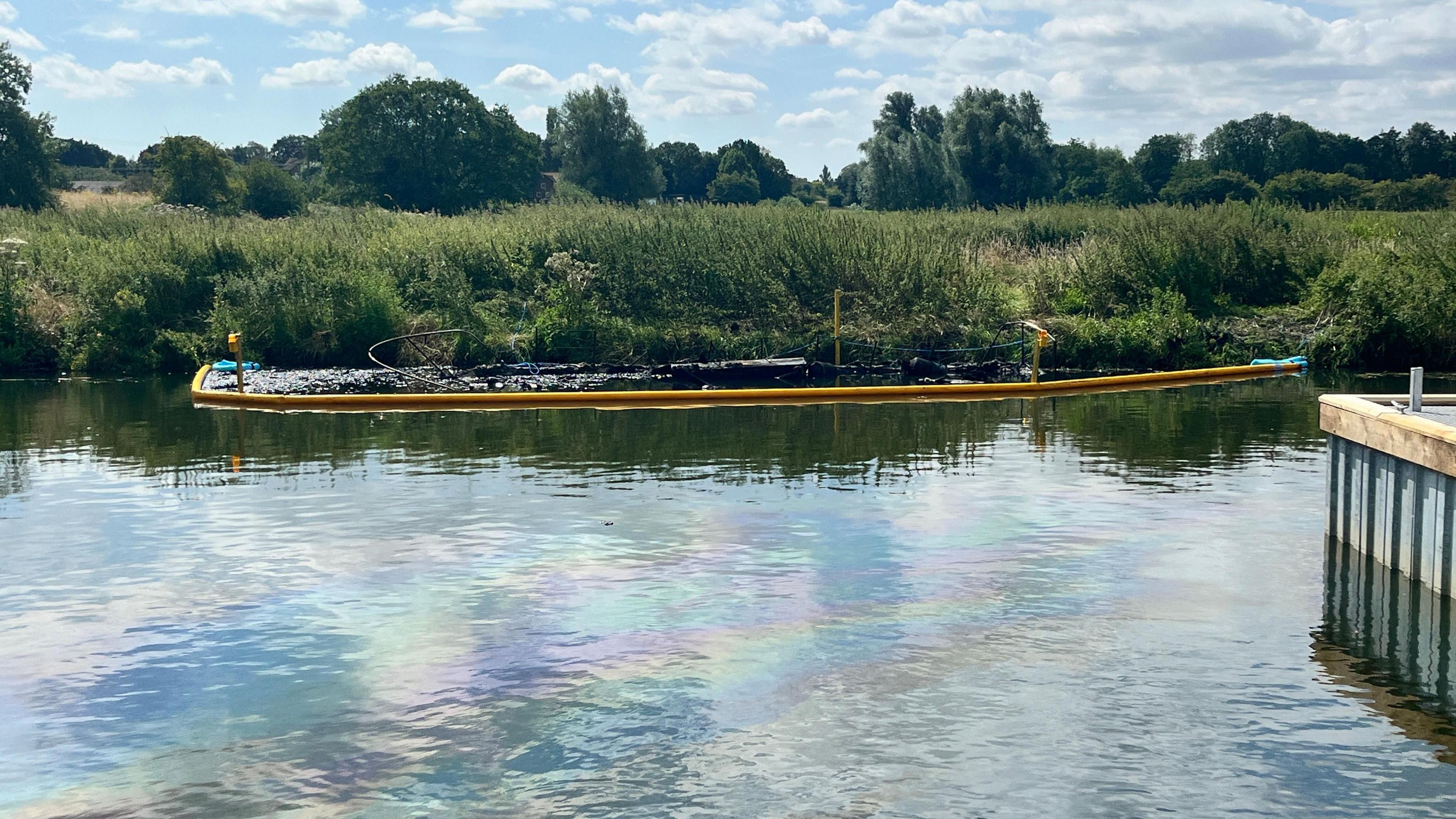 A burnt-out boat by a river bank. An oil sheen can be seen on the water. Beyond the bank is a field and trees.