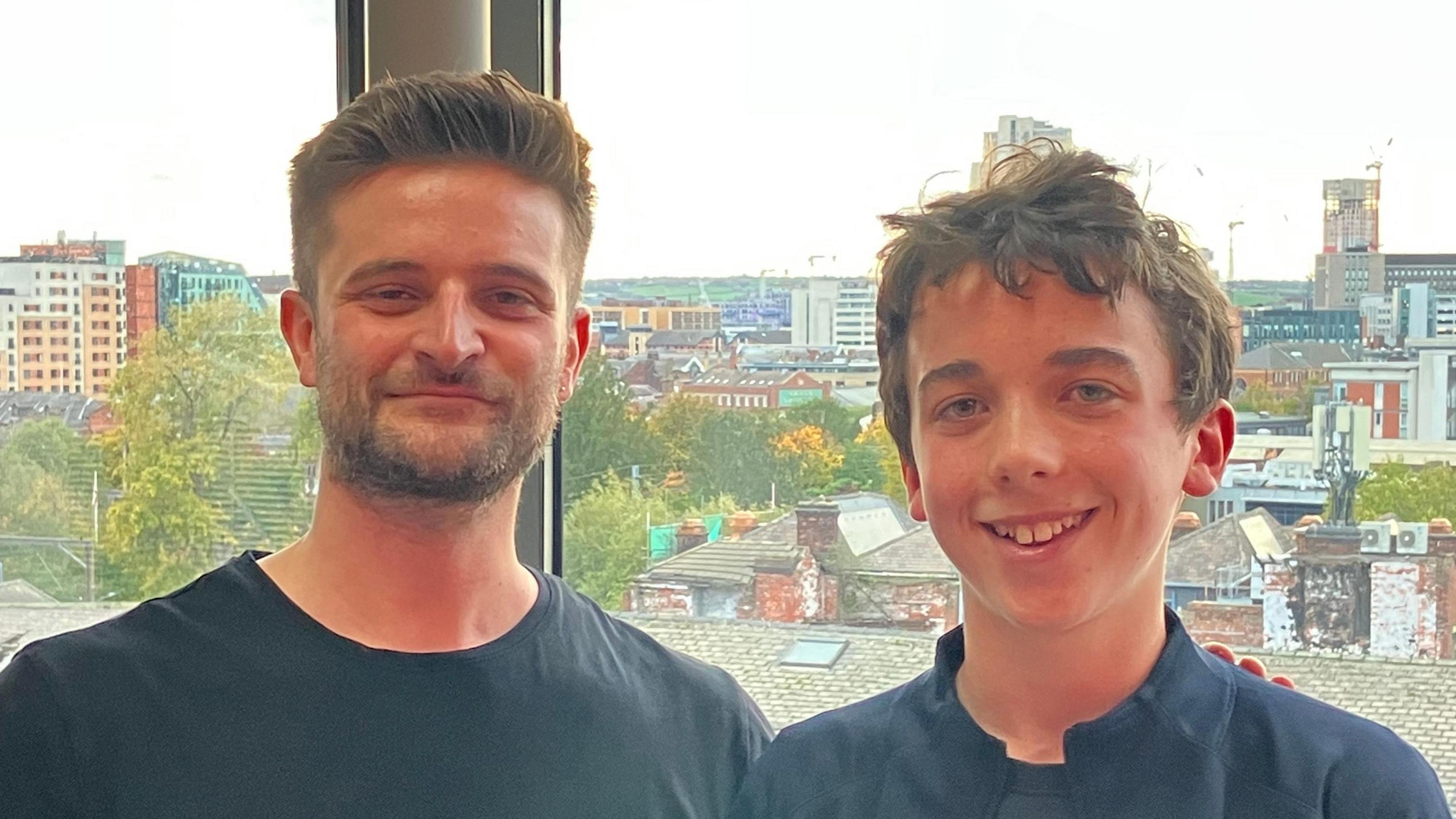 Barnaby and Nic are standing indoors in front of a large window with an urban cityscape visible in the background. The view outside includes a mix of modern and older buildings, rooftops, and trees with autumn foliage. Construction cranes and tall structures can be seen in the distance under a bright sky. Both are wearing dark tops and are positioned close together, with one person’s arm resting on the other’s shoulder.