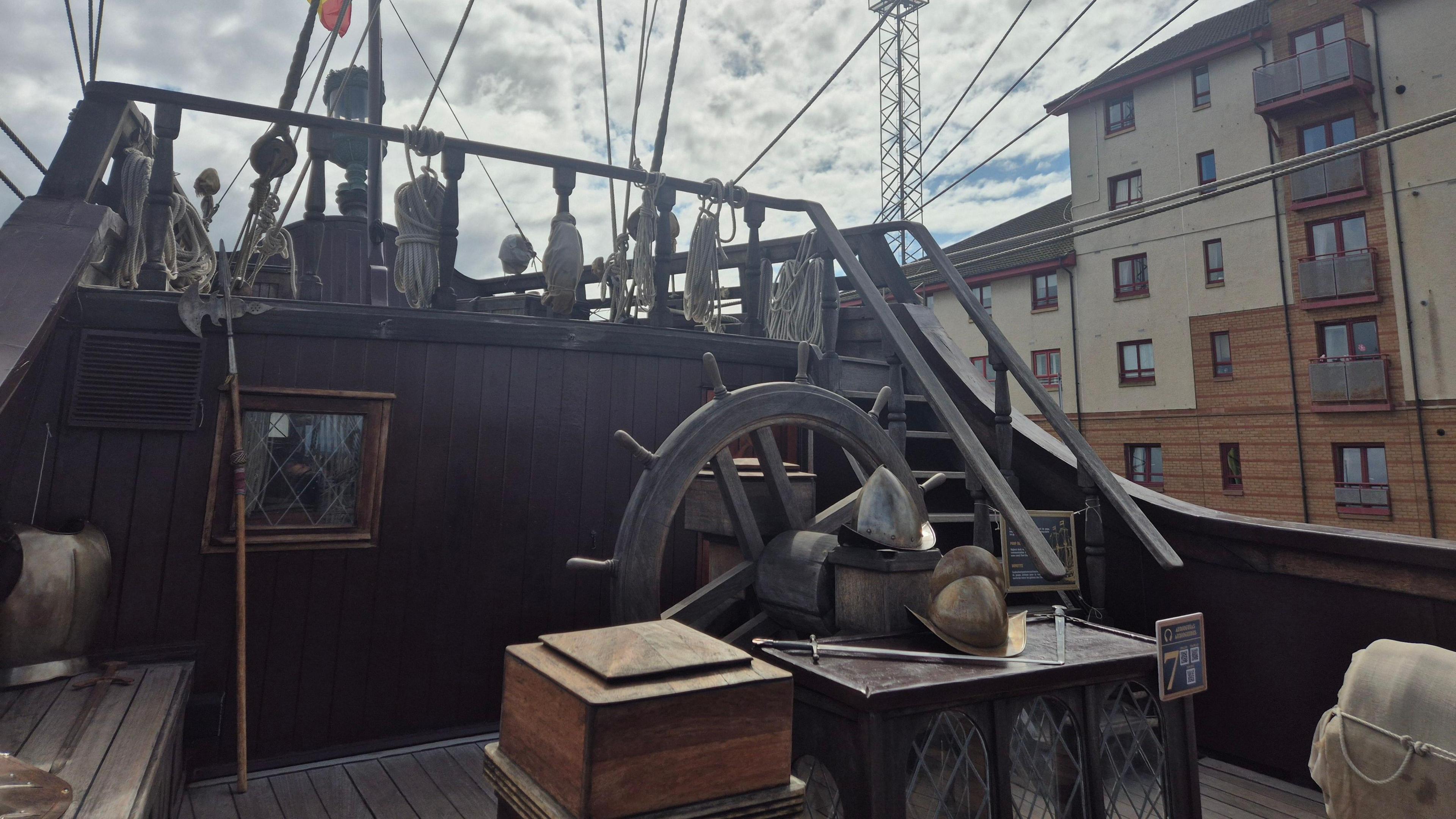 An elevated wooden deck area with extensive rigging lines stretching upward and outward, and a large traditional ship's wheel positioned near the centre.